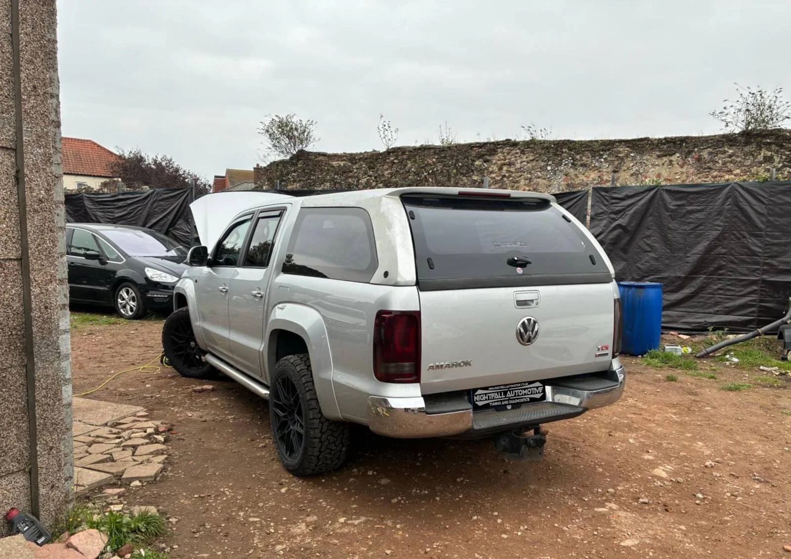 Silver Volkswagen Amarok pickup truck with a canopy, parked on a dirt lot, with a black wheel on the rear and a trailer hitch.