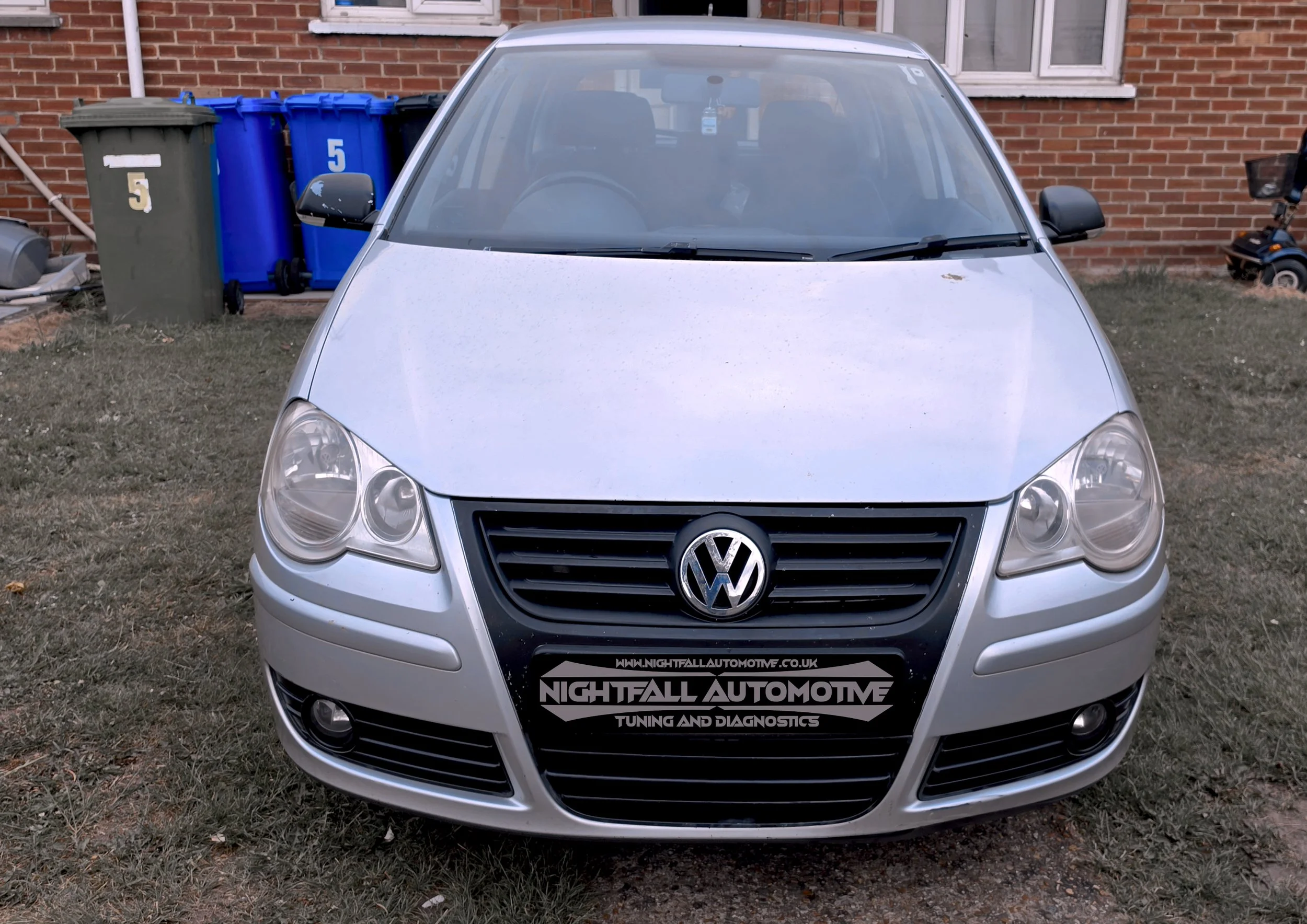 Front view of a silver Volkswagen car parked on grass in front of a brick house with blue and grey wheelie bins nearby. tuned by Nightfall Automotive