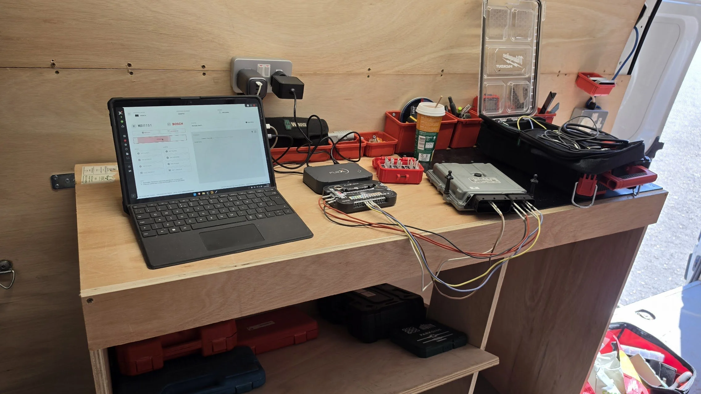 A workbench with electronic tools and equipment, including a tablet displaying a diagnostic interface, various wires and circuit boards, a red toolbox, and a black carrying case for tools, set against a wooden wall.