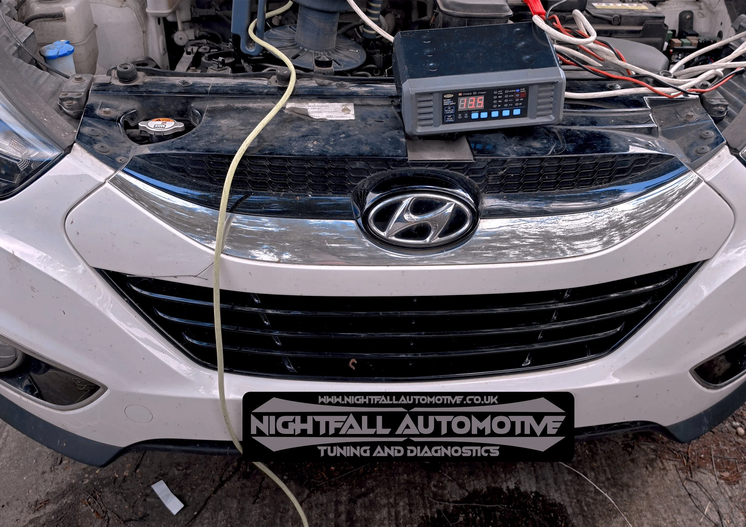 A car with its hood open undergoing tuning and diagnostics, with a battery charger connected and a display showing voltage levels, parked on dirt ground.