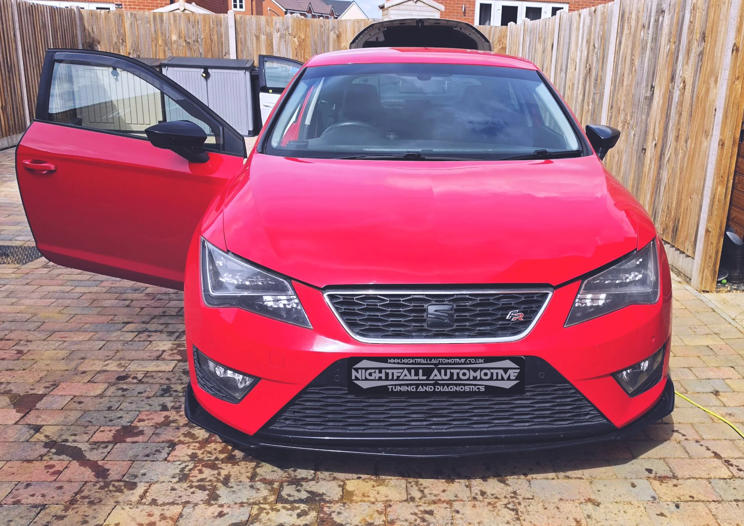 Red hatchback car with a black front grille, parked on a brick driveway, with the driver's door open and a fence in the background. Nightfall Automotive