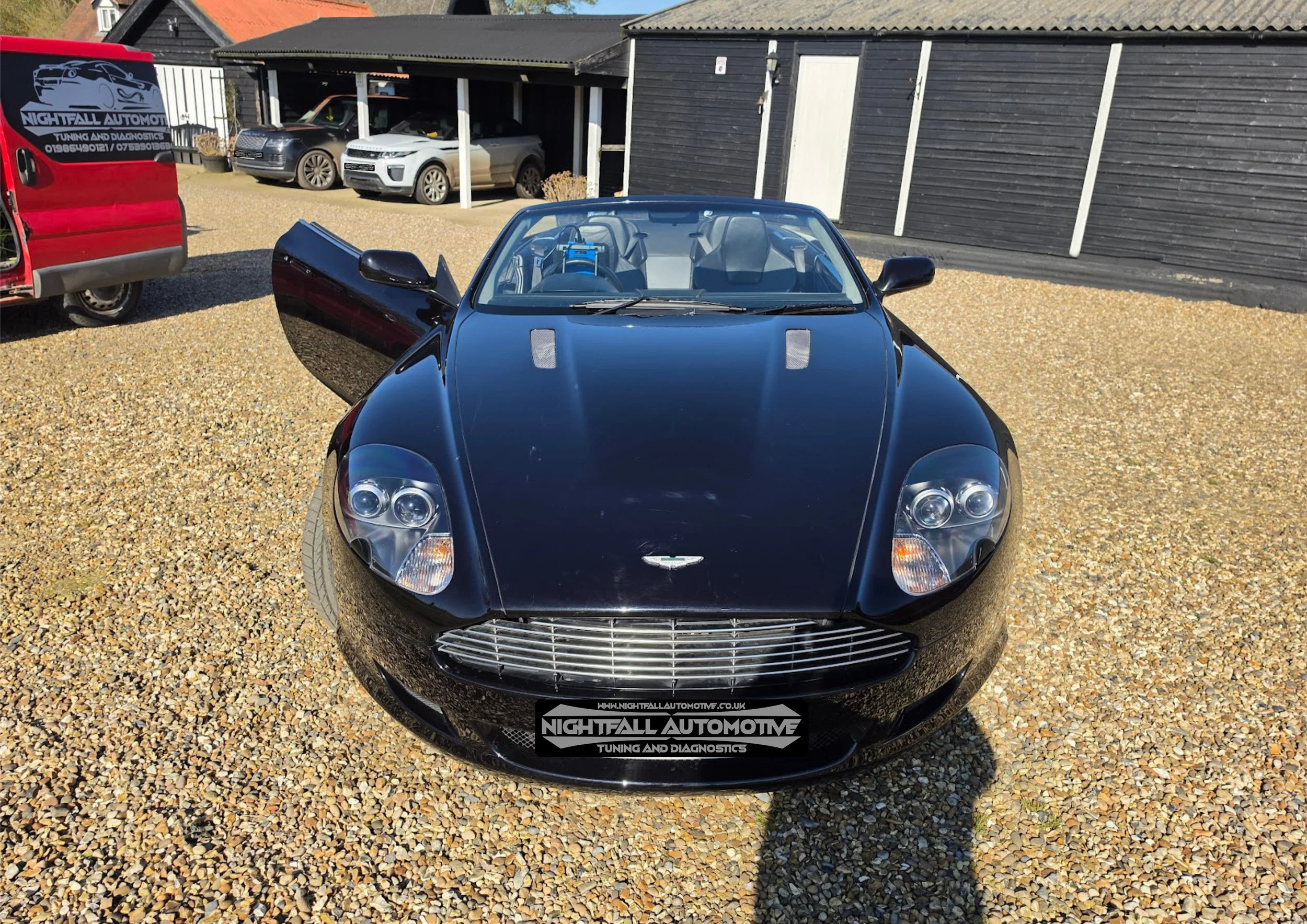 Black convertible sports car parked on gravel with one door open, at an automotive tuning and diagnostics shop. Several other cars and a red van are visible in the background, along with a black building.