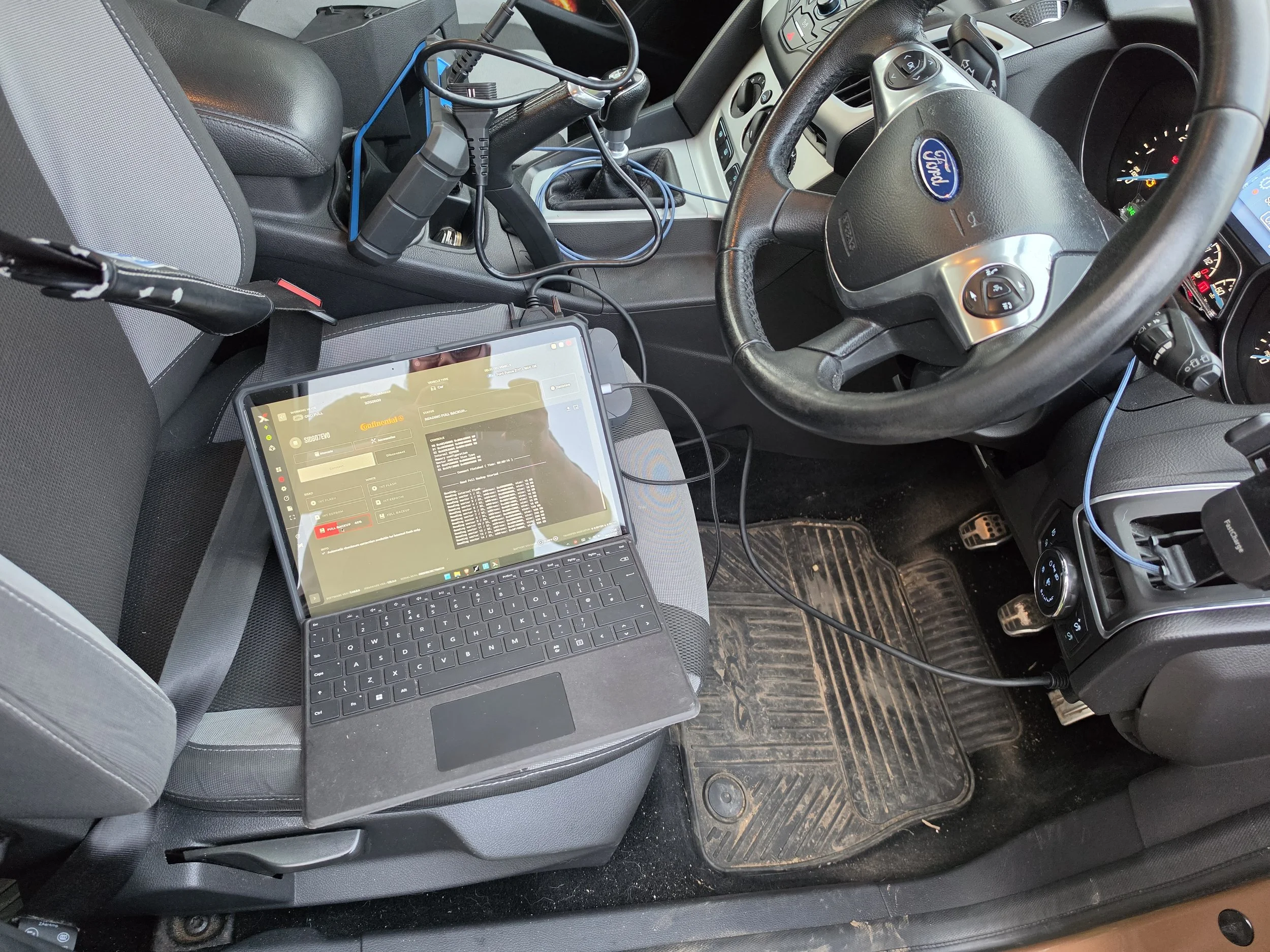 Inside a vehicle with a laptop on the passenger seat connected to various wires and equipment, including a hand-held device, with a dirty floor mat and the steering wheel visible.