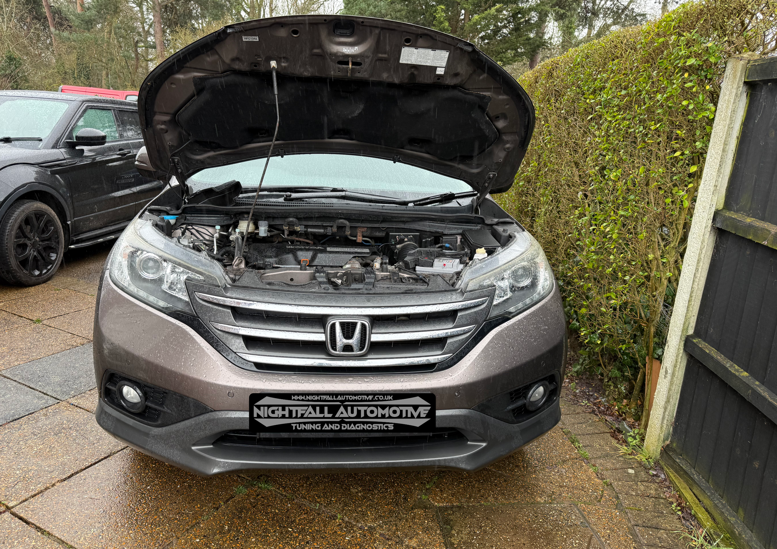 A silver Honda SUV with its hood open, parked on a wet driveway bordered by a black fence and green bushes. A second black car is parked next to it.