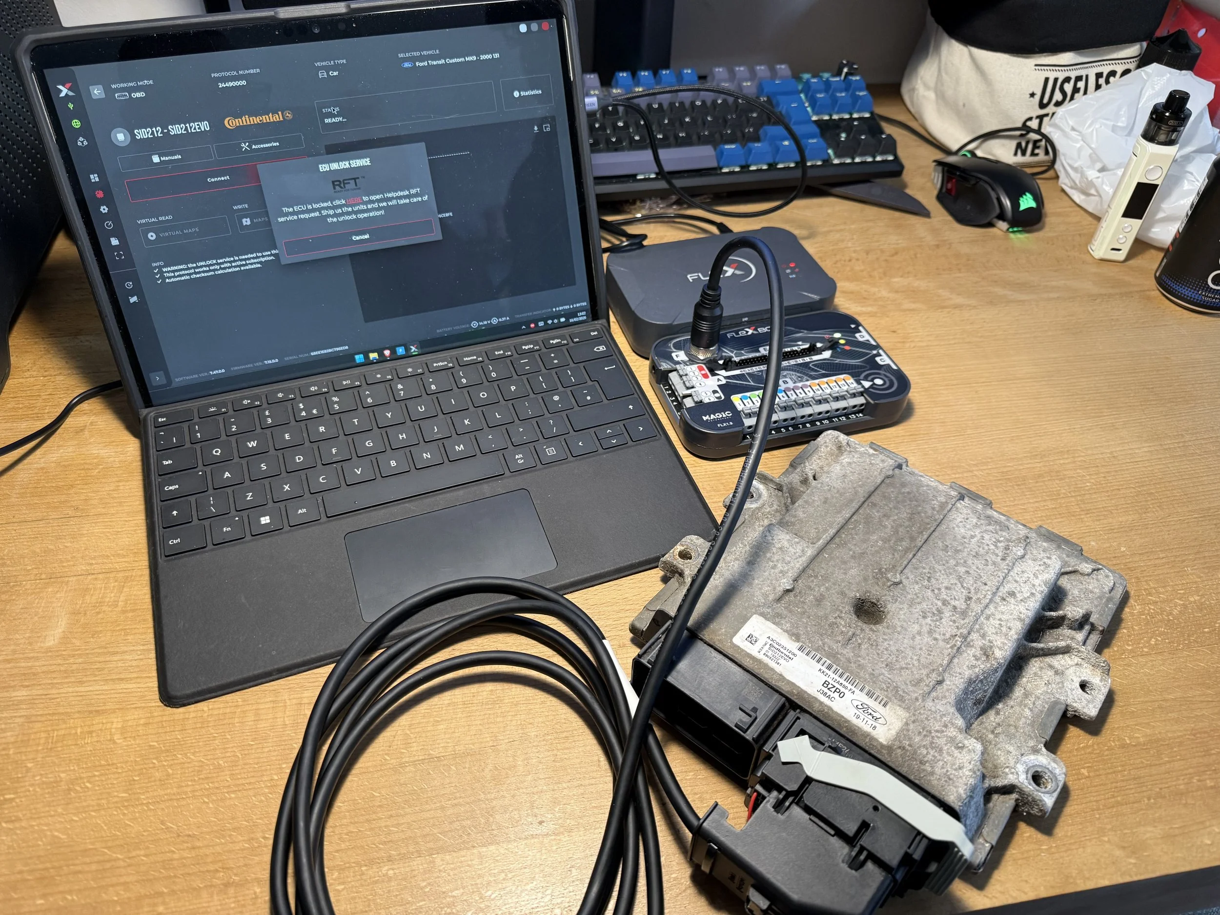 A workspace with a laptop, an ECU, and various electronic devices, including a keyboard, cables, and a vape pen on a wooden desk.