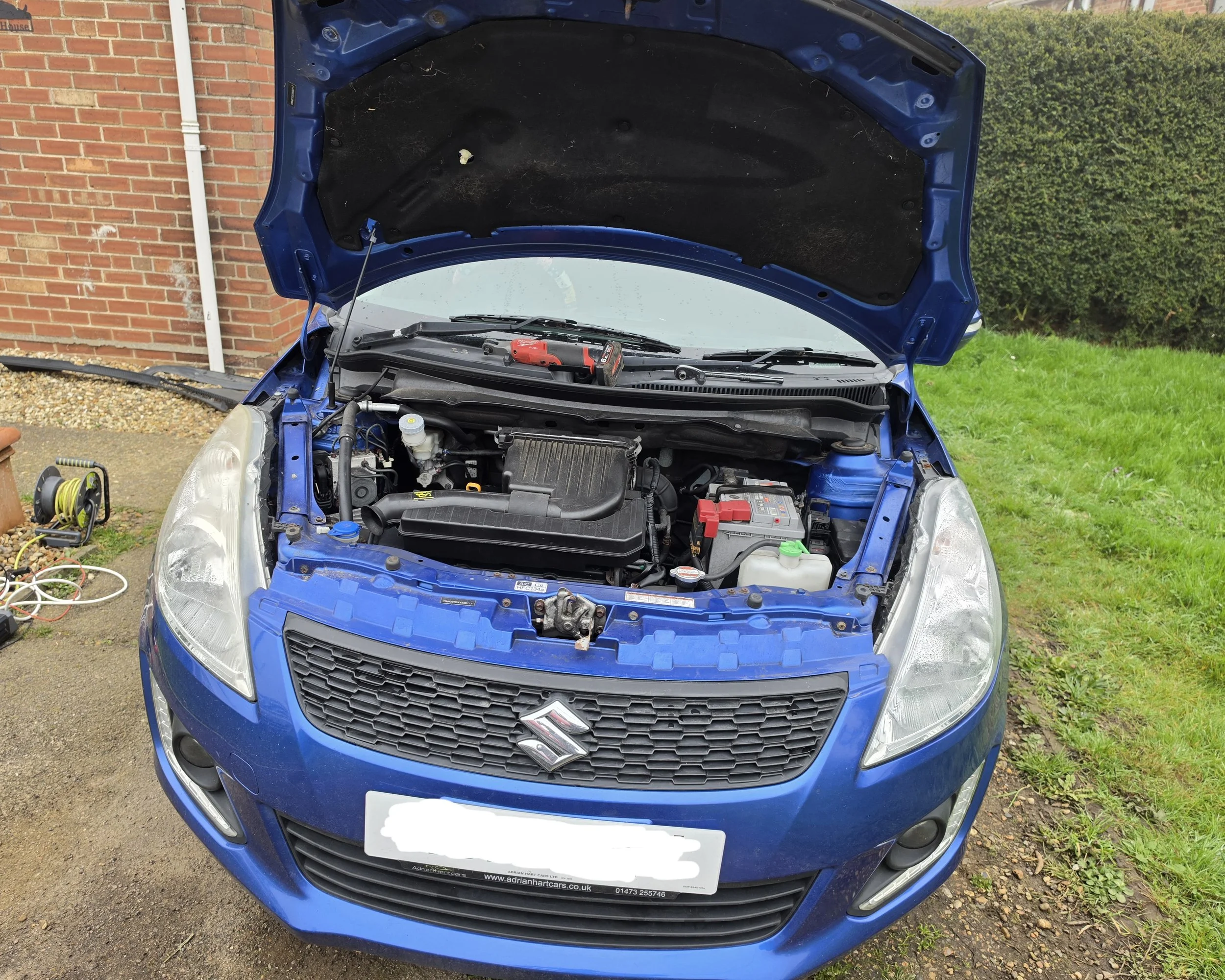 Blue car with open hood showing engine bay, outside near a brick building and a lawn, with tools and equipment nearby for car maintenance.