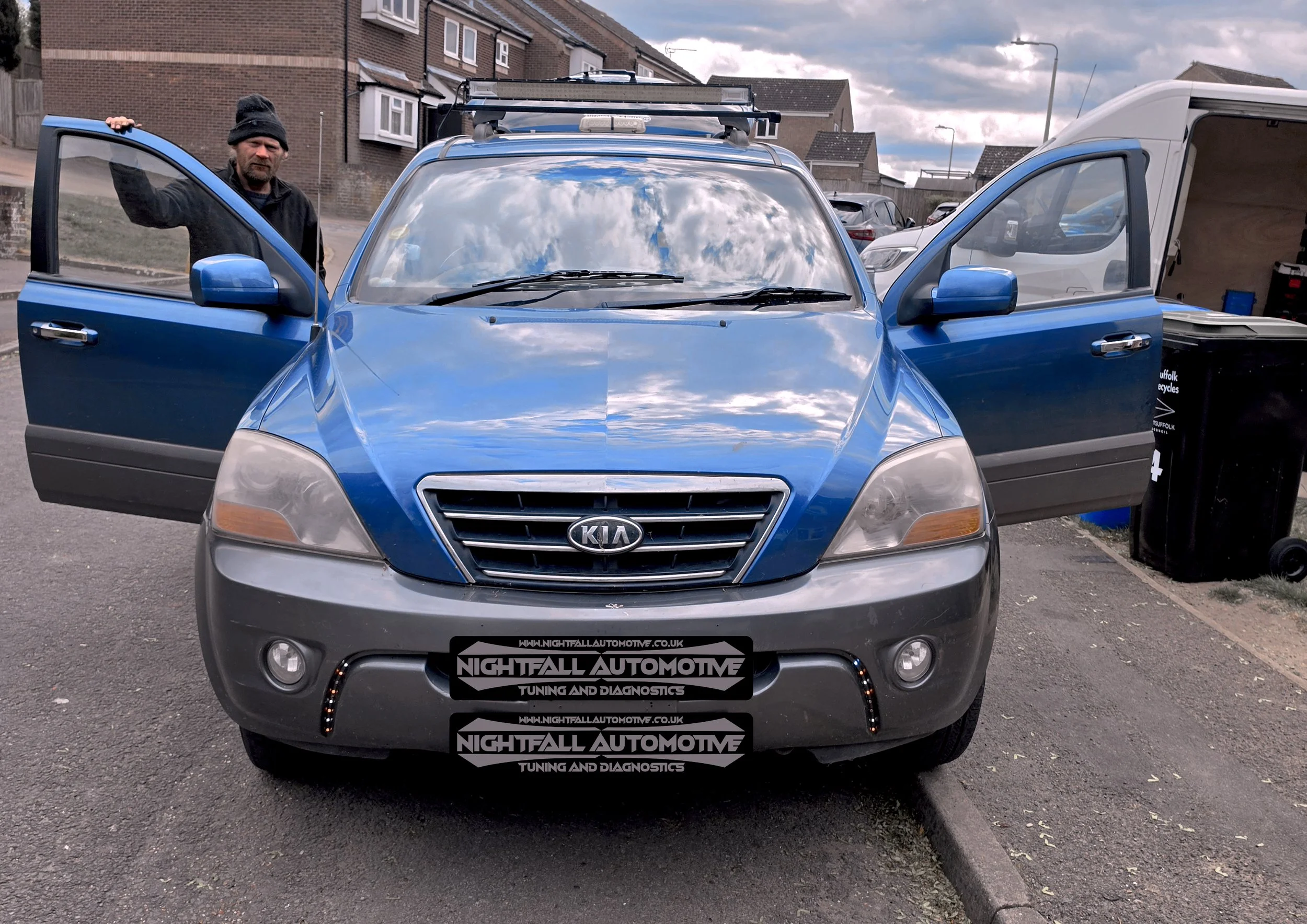A man wearing a black beanie and black jacket standing next to a blue Kia Soul car with open driver’s side door. Tuned by Nightfall Automotive
