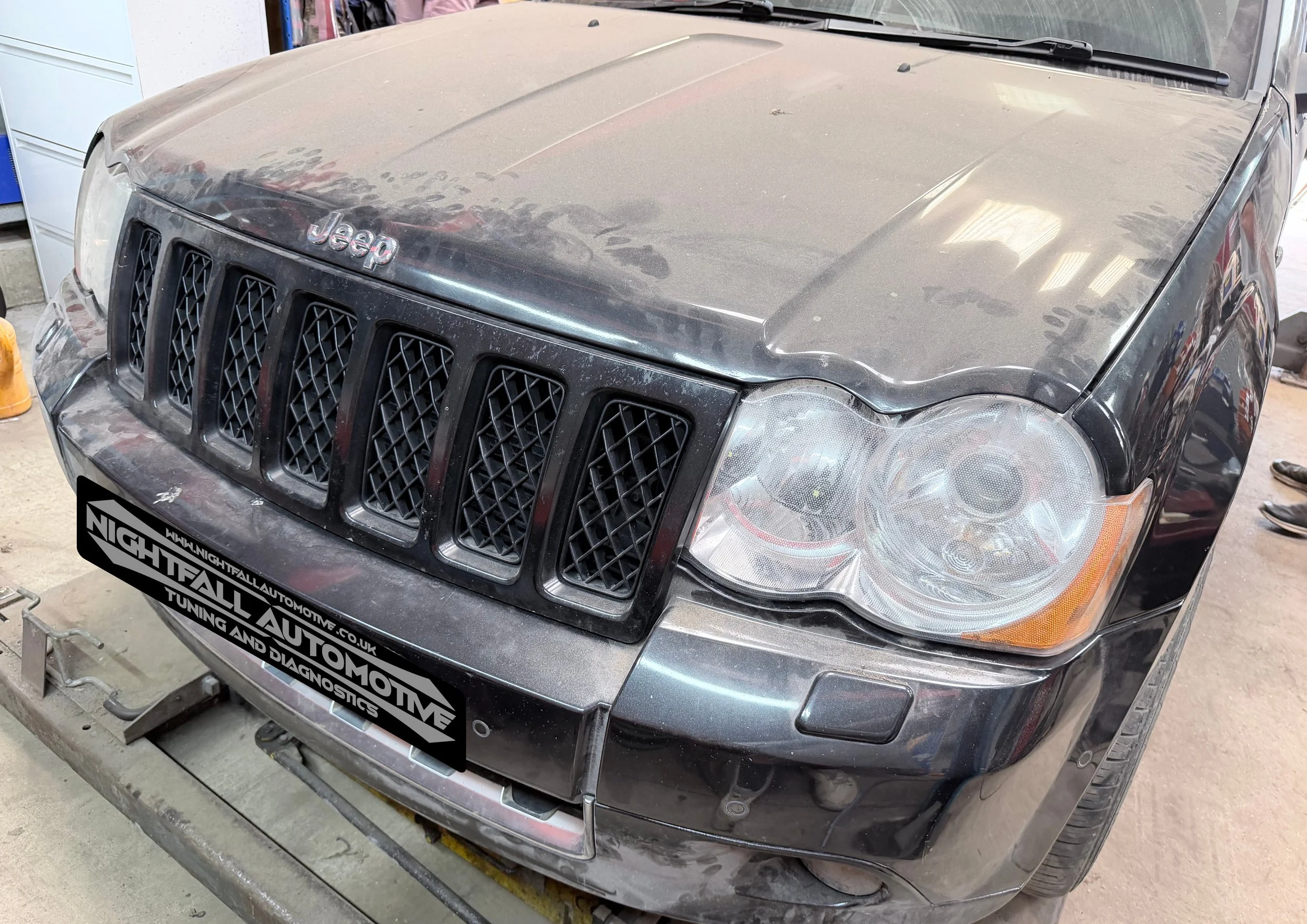 A dusty black Jeep Grand Cherokee with a damaged front bumper and a license plate that reads 'NIGHTFALL AUTOMOTIVE.' The vehicle is inside a garage, surrounded by various tools and equipment. Tuned by Nightfall Automotive