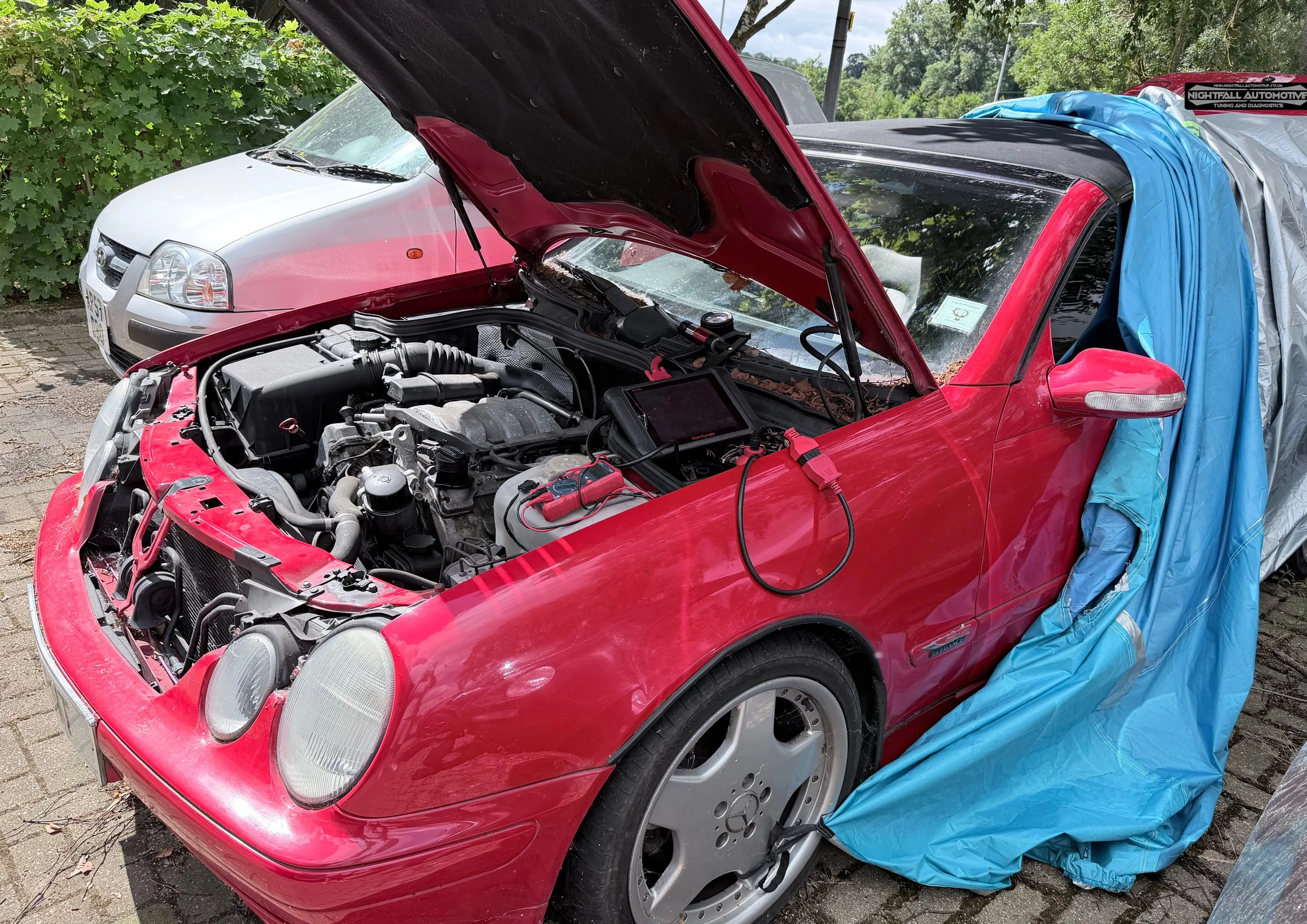 A red sports car with its hood open, revealing the engine, parked next to a white sedan. Blue and gray tarps are draped over part of the red car. The scene is outdoors, near green trees and a cloudy sky.