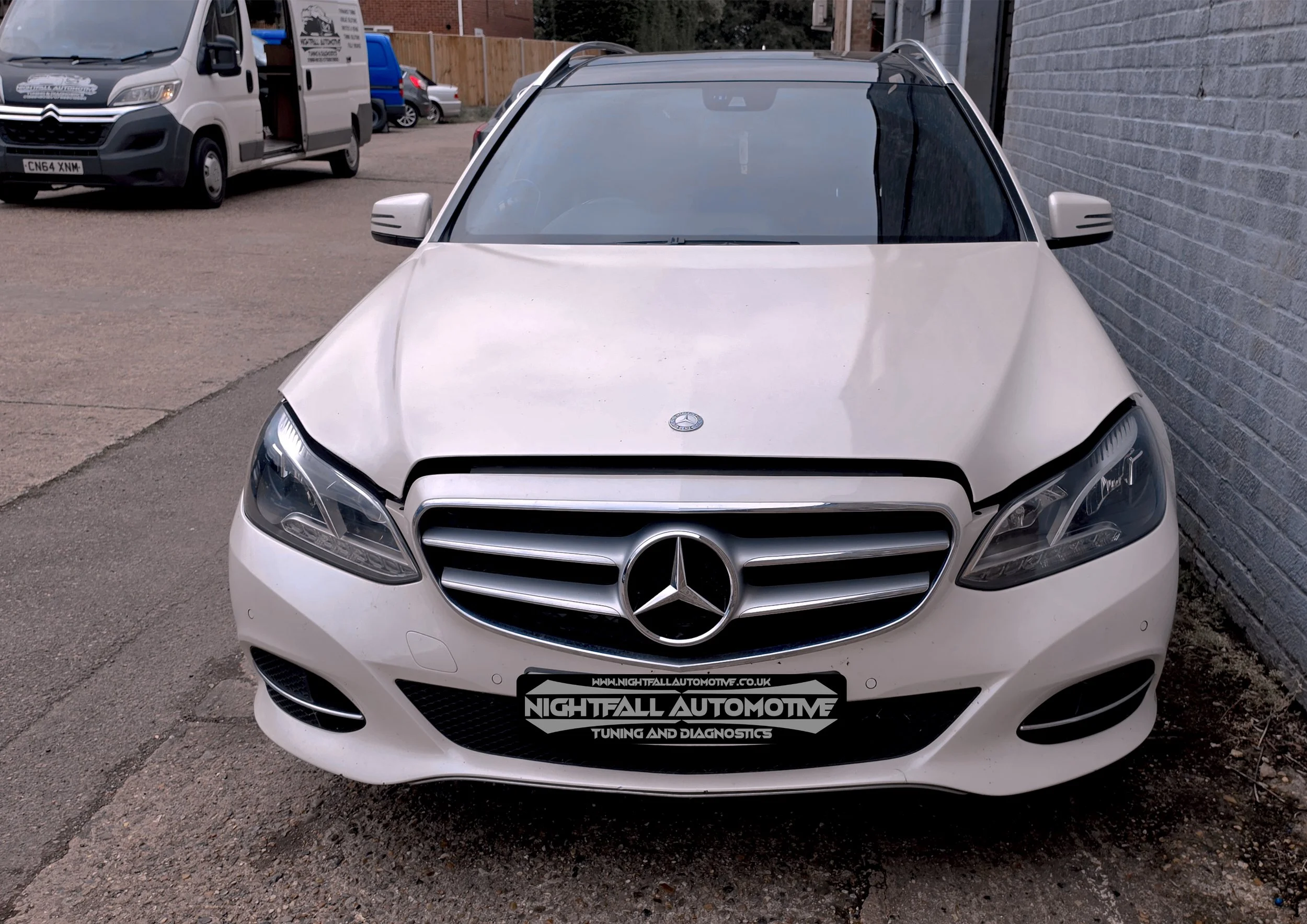 Front view of a white Mercedes-Benz vehicle parked next to a brick wall with a logo for Nightfall Automotive on the front license plate. Other vehicles are visible in the background, including a white van and a blue car.