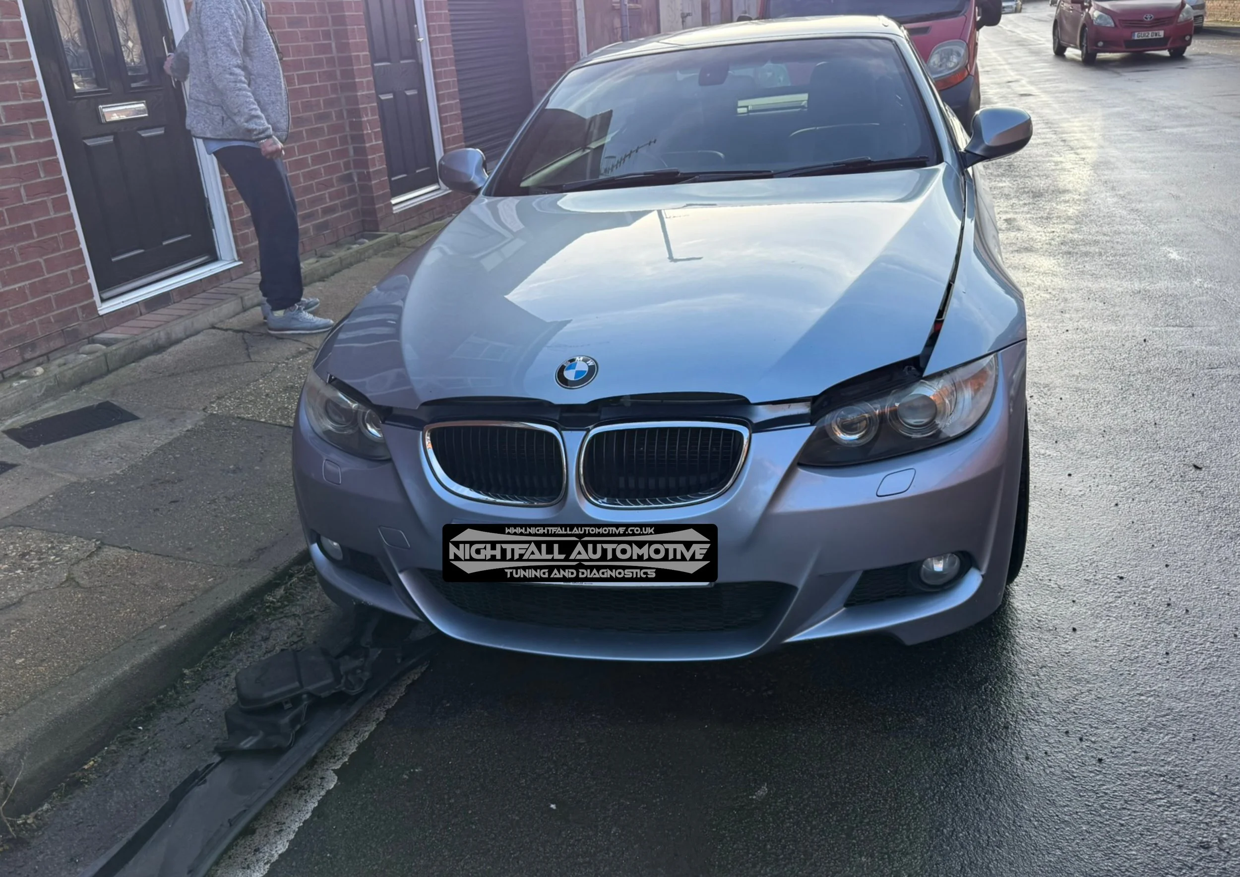 A silver BMW car parked on the side of the road with front-end damage, including a bent hood and broken headlight, in front of a brick house with a person standing nearby.