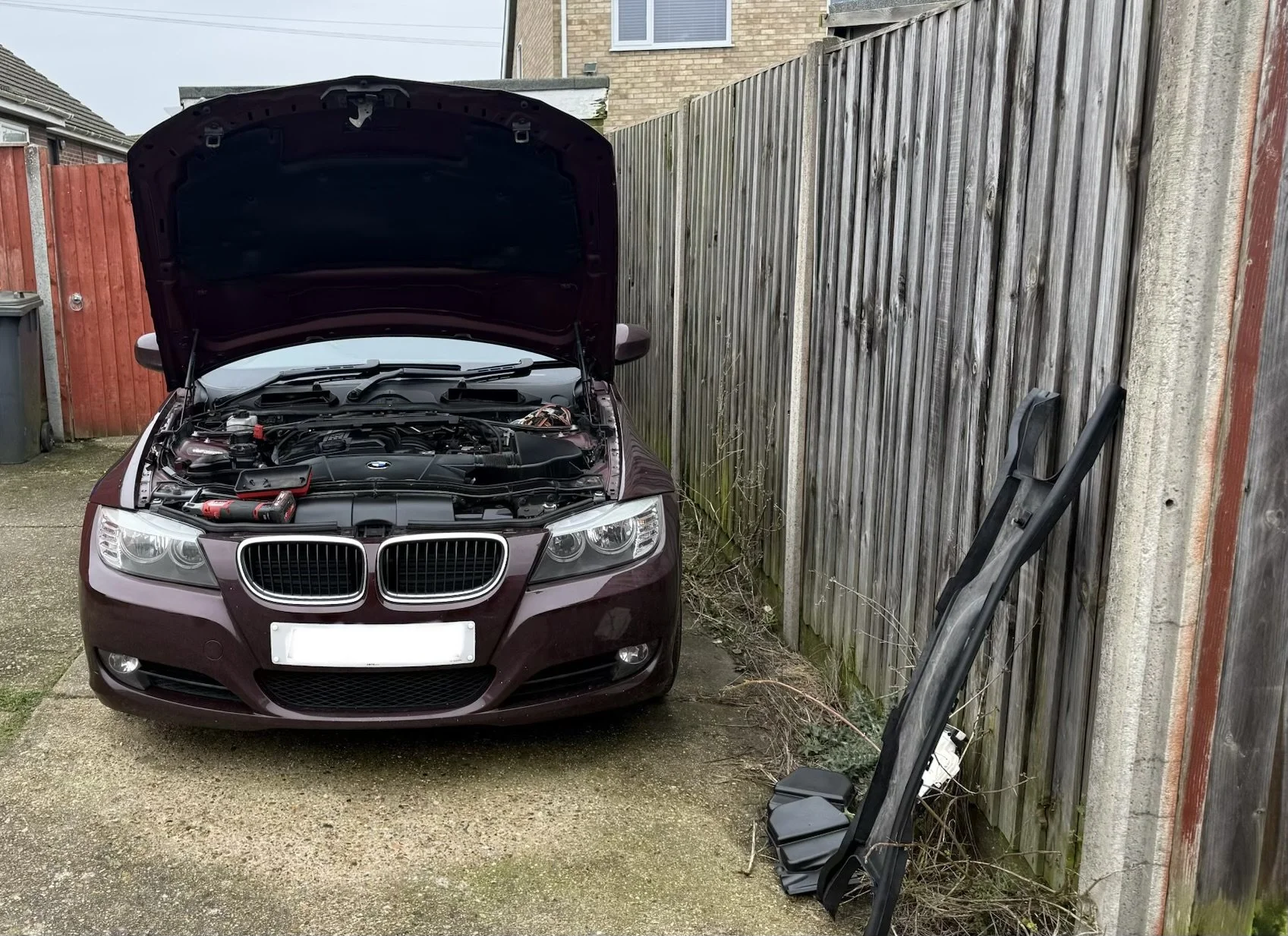 A maroon BMW car with open hood parked in a driveway next to a wooden fence, with tools and a car part leaning against the fence. Stage 1 by Nightfall Automotive
