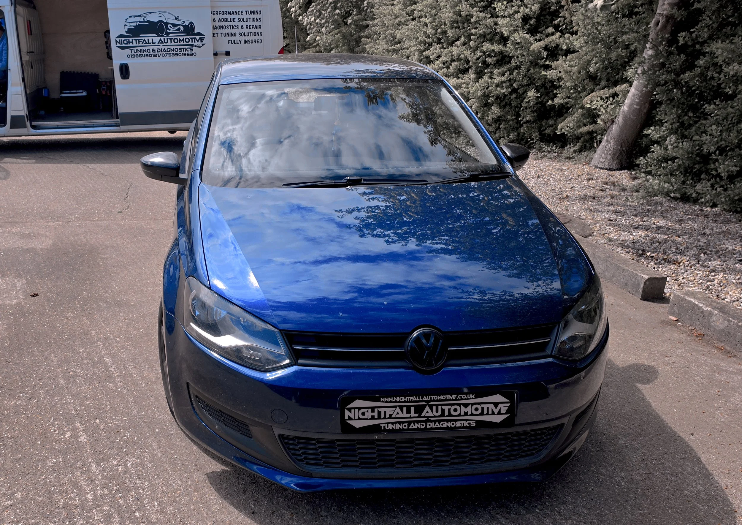 Blue Volkswagen car parked in a parking lot with a backdrop of bushes and trees, and a white commercial van with a logo and text related to automotive tuning and diagnostics. Tuned by Nightfall Automotive