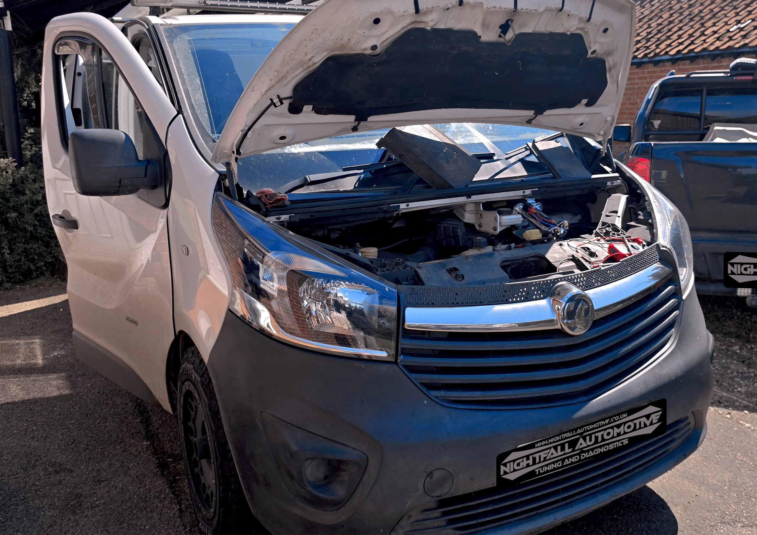 A silver pickup truck with its hood open, revealing the engine and internal components. The truck is parked outside with other vehicles and a building with a brick wall and tiled roof in the background. There is a sticker on the front bumper for Nigh