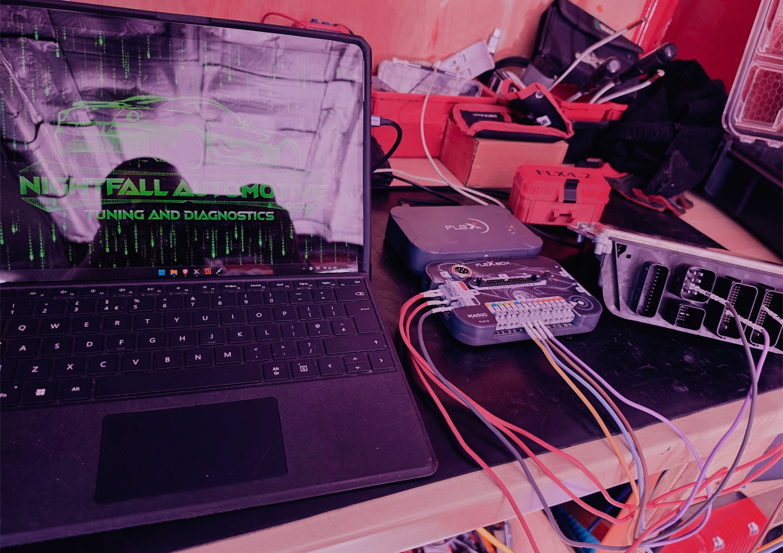 Workstation with a laptop displaying code and a digital car, connected to automotive diagnostic tools and equipment on a cluttered workbench. stage 1 by Nightfall Automotive