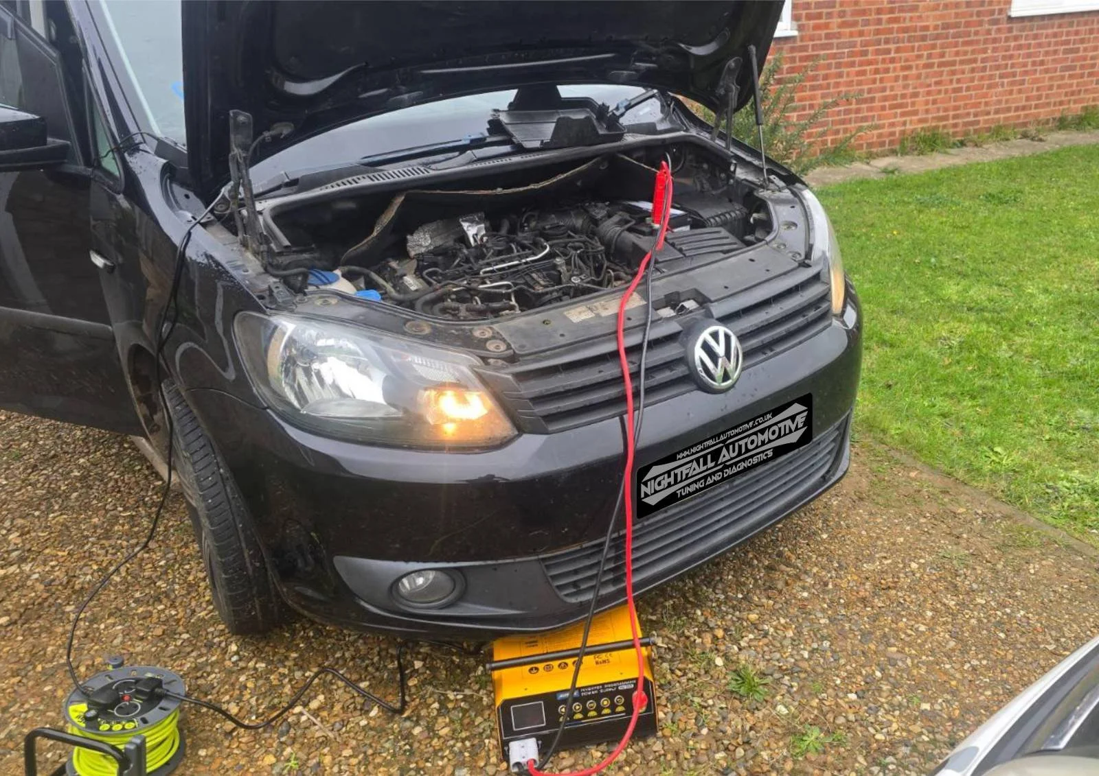 Black Volkswagen car with its hood open, connected to testing equipment on a gravel driveway near a brick wall, with a patch of green grass nearby.