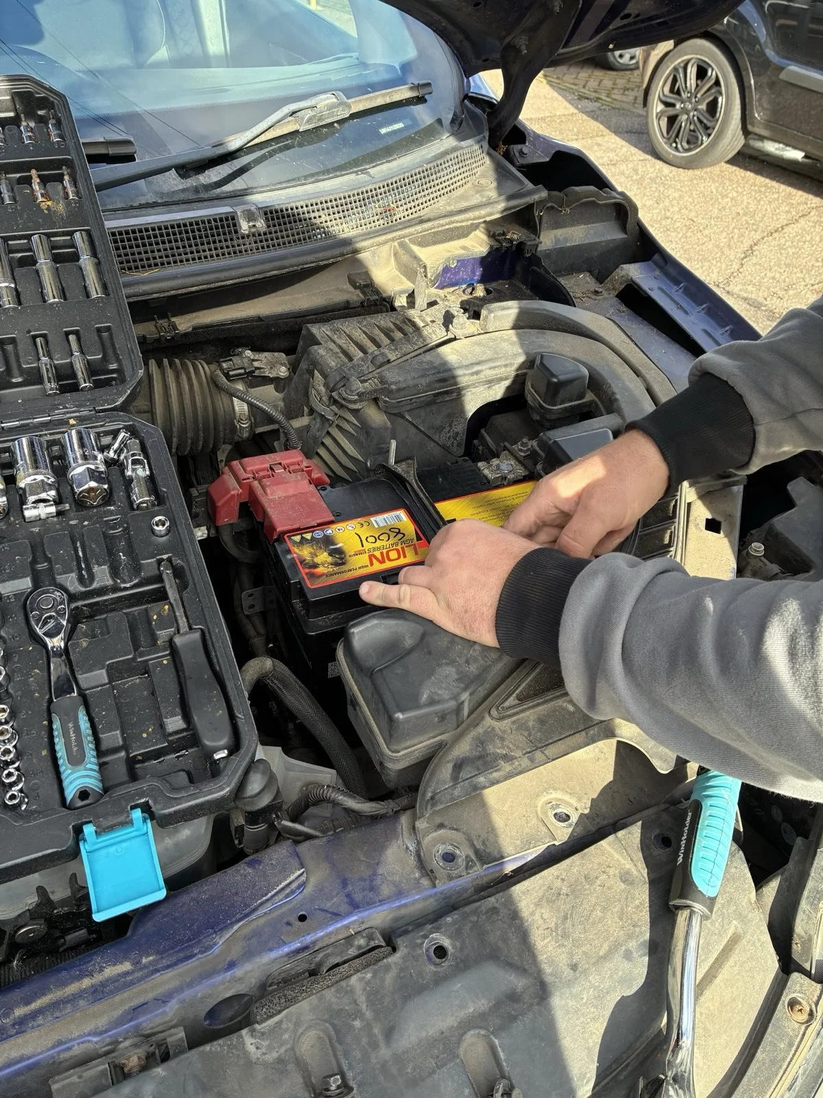 A person installing a car battery in an engine bay with tools nearby.