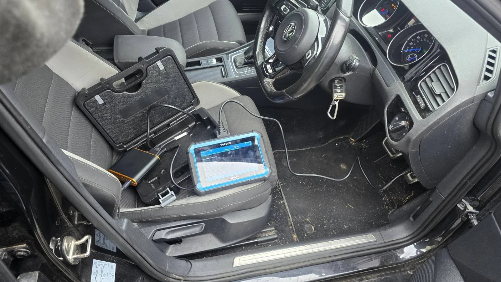 The interior of a car with a diagnostic scanner and portable electronic devices placed on the passenger seat, connected with cables.