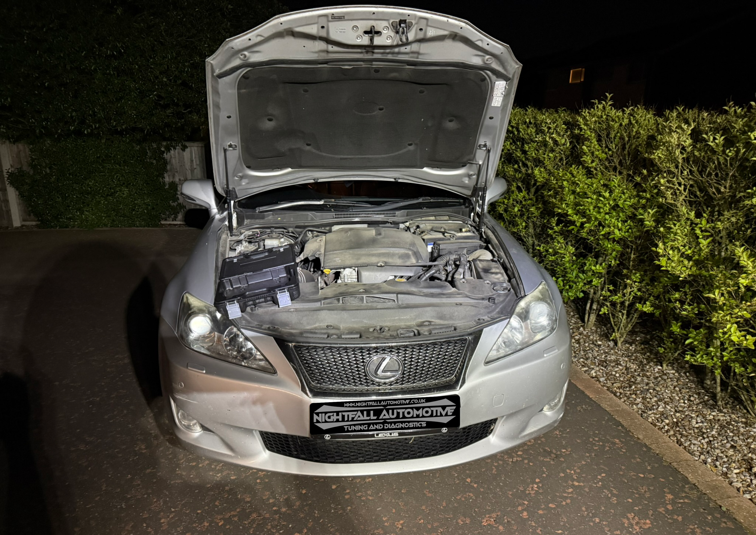A silver Lexus car with its hood open, parked on a driveway at night, with bushes and trees in the background.