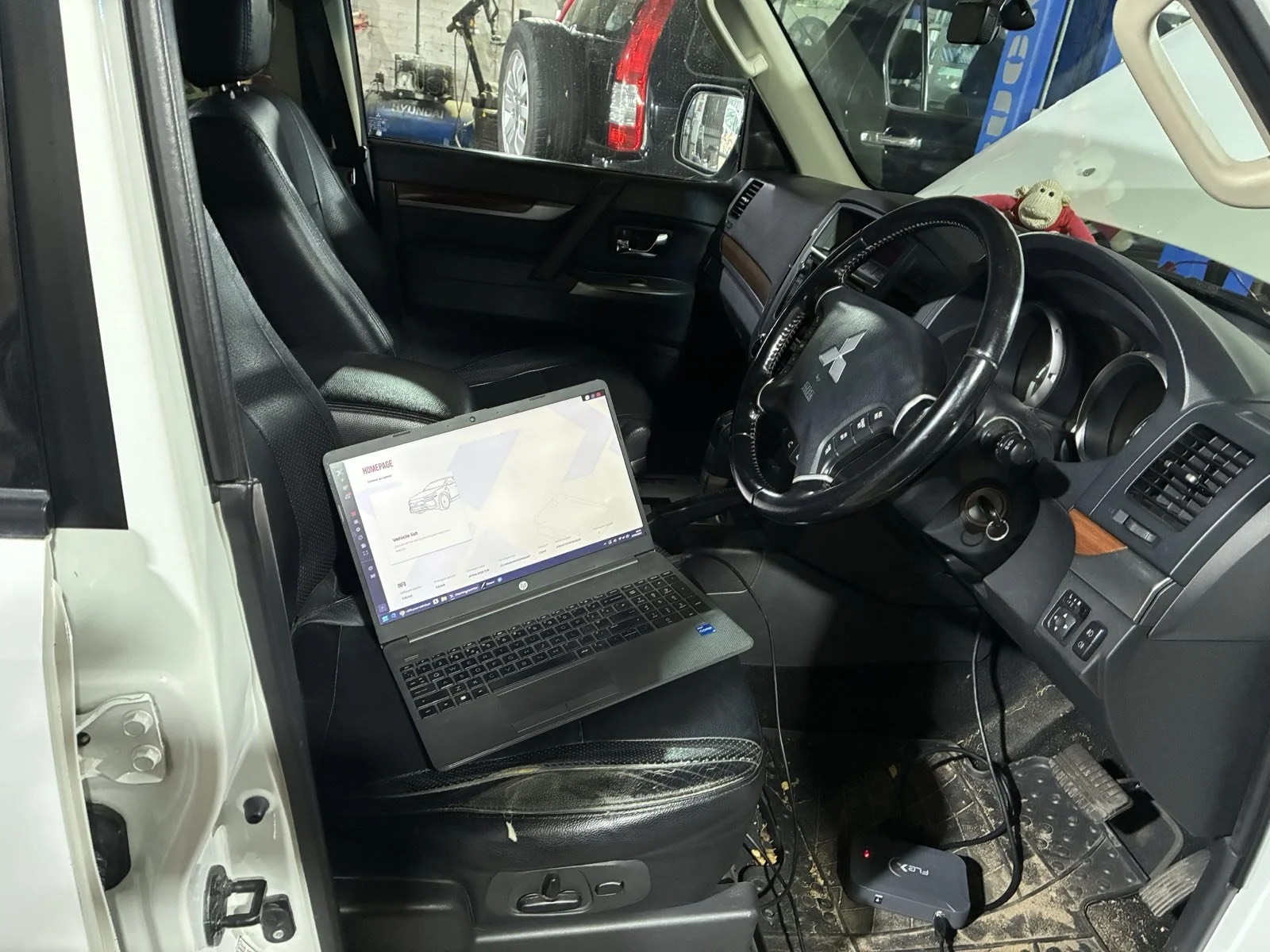 Interior of a vehicle with a laptop on the driver's seat, a stuffed monkey on the dashboard, and windshield view showing other parked vehicles.