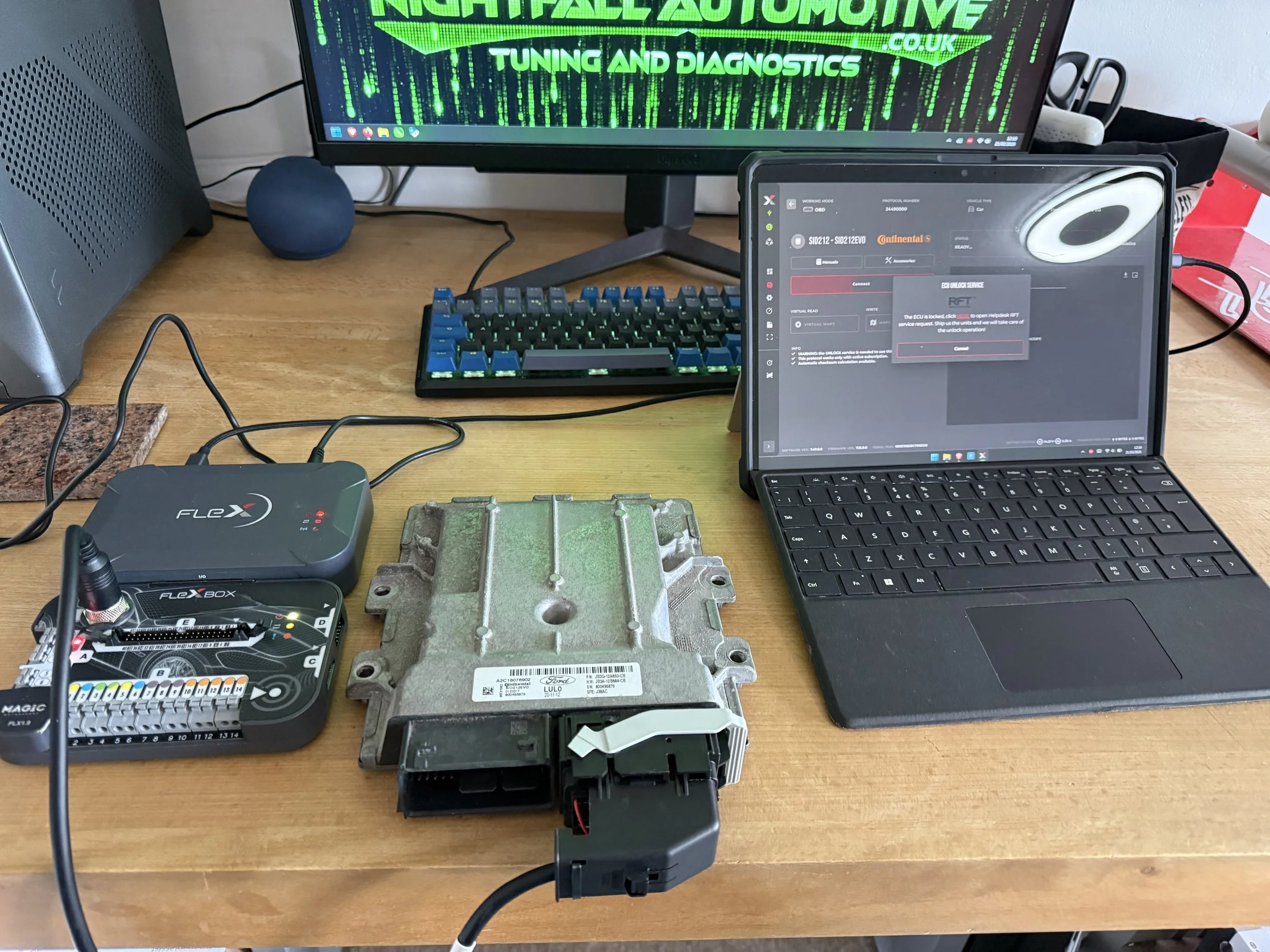 A computer workspace with a car engine control module, a diagnostic device, a small all-in-one computer, a monitor, a keyboard, and a small speaker on a wooden desk.