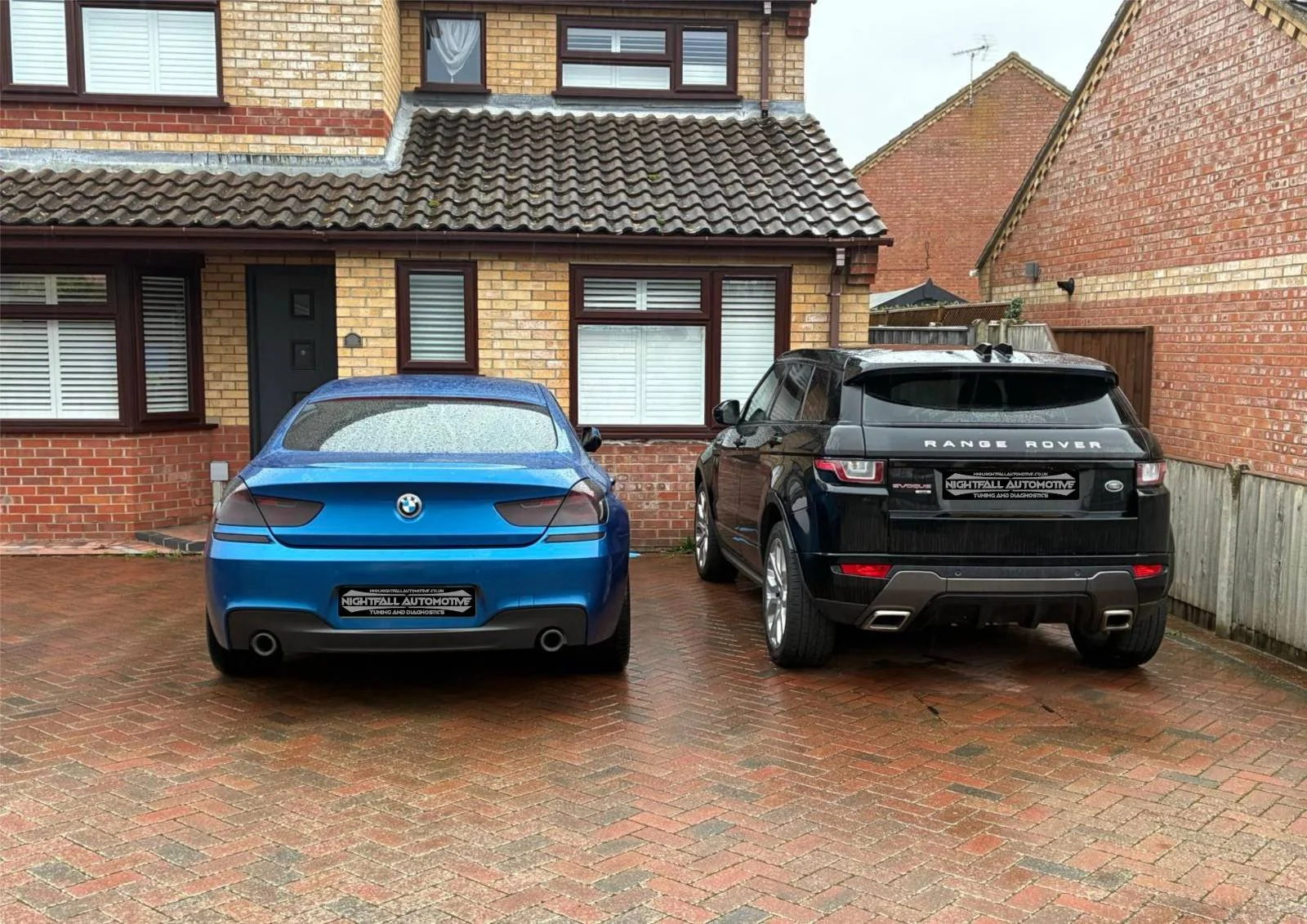 A blue BMW and a black Range Rover parked in front of a brick house with rain droplets on the BMW