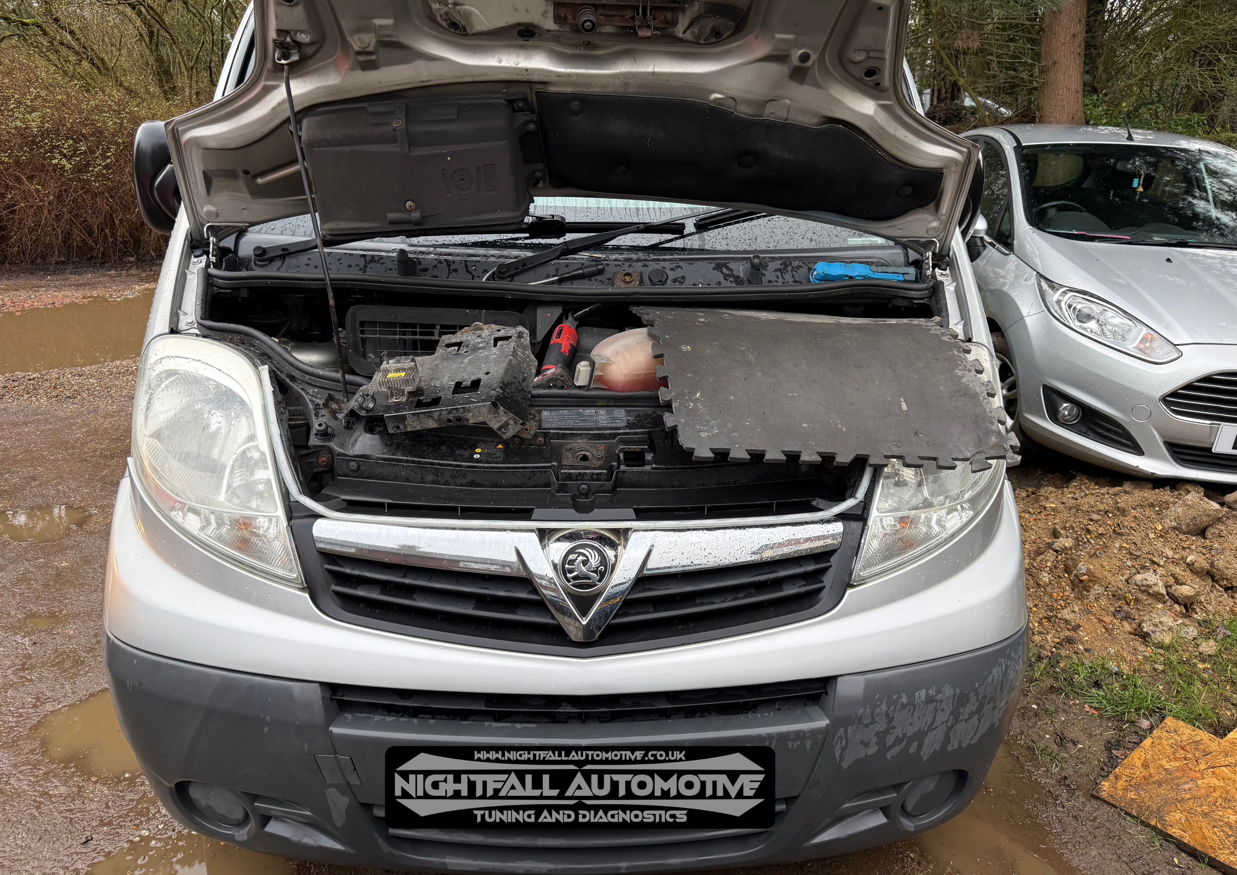 Front view of a silver Vauxhall vehicle with its hood open, revealing the engine bay, parked on muddy ground. A black and white sign with the text 'Nightfall Automotive Tuning and Diagnostics' is attached to the front bumper. Next to it is a silver c