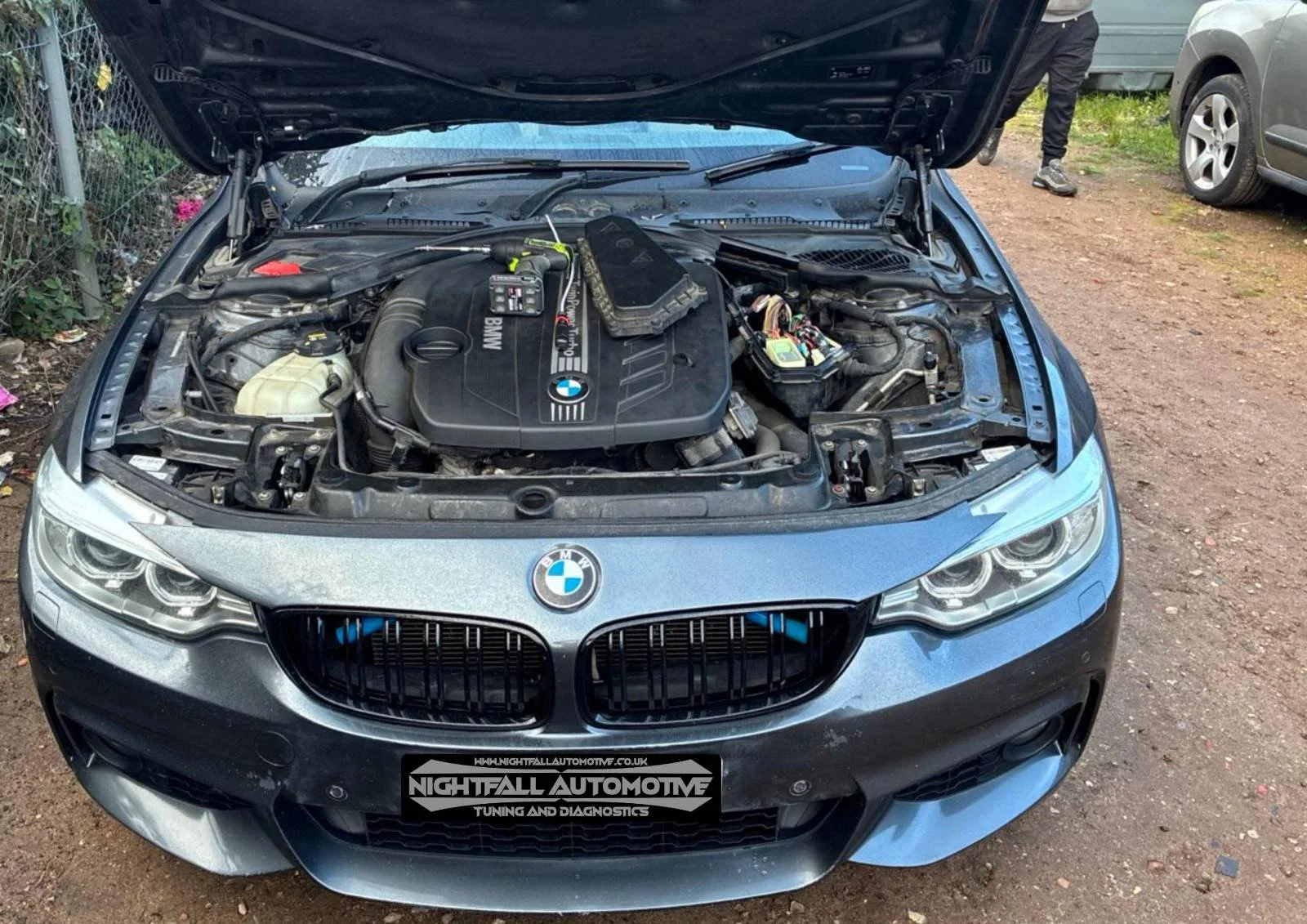 Open hood of a BMW car showing its engine and internal components, parked outdoors on a gravel surface.