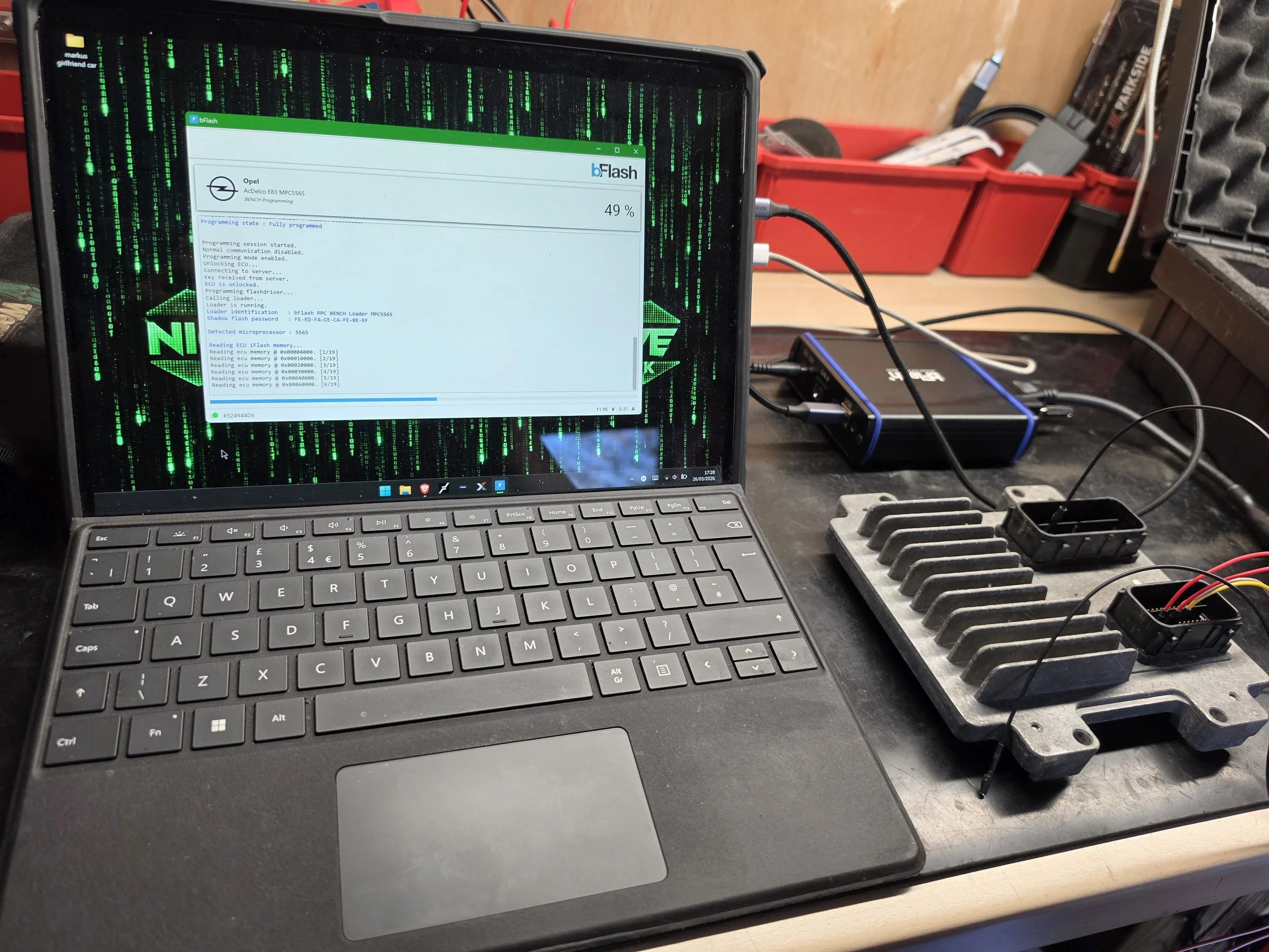 A laptop with a touchscreen displaying a programming interface for flashing an Opel vehicle's electronic control unit (ECU). The laptop is on a workbench alongside tools and electronic components, with a red tool organizer and various cables in the b