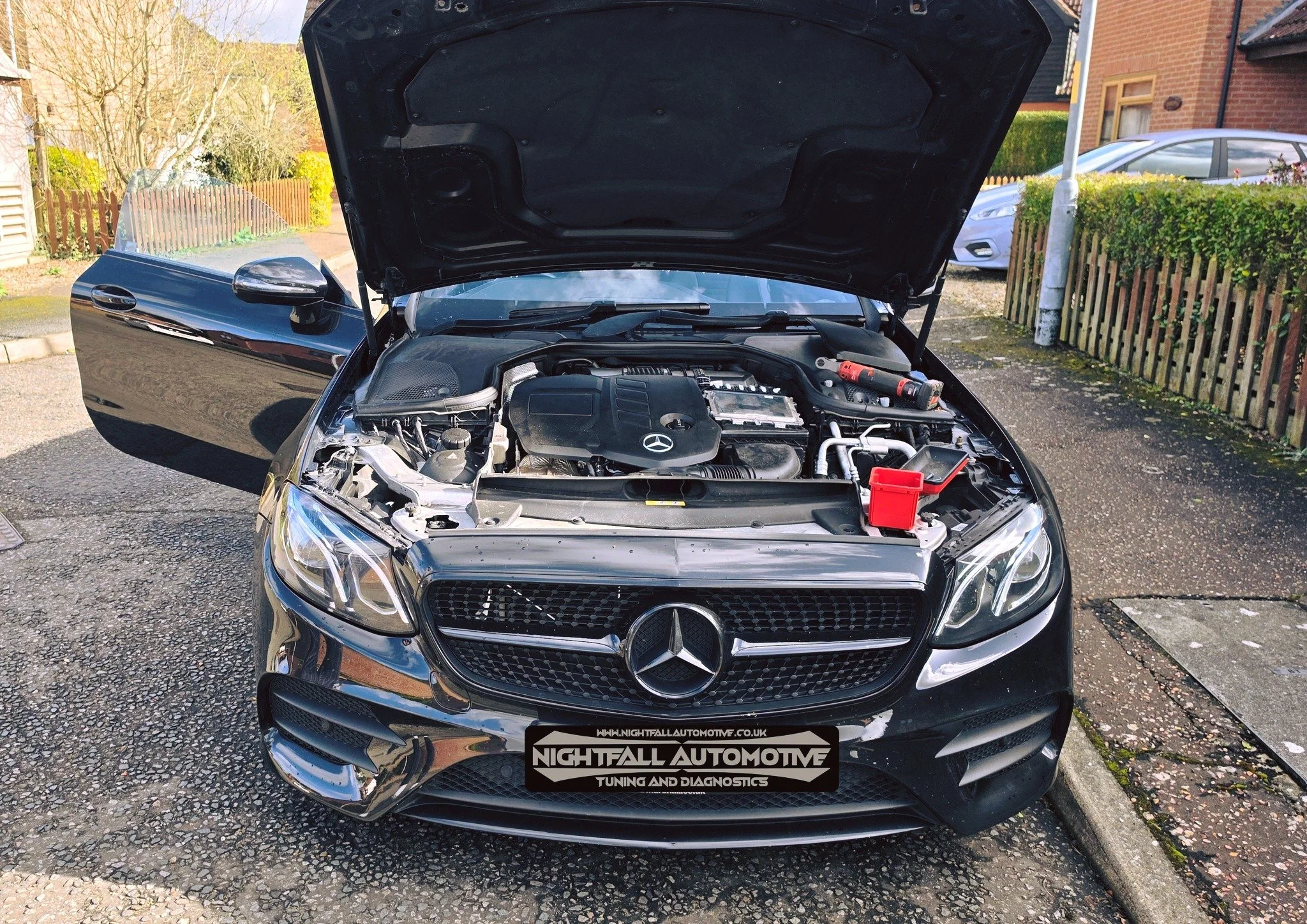 Front view of a black Mercedes-Benz car with its hood open, parked on a street with a residential area and trees in the background.