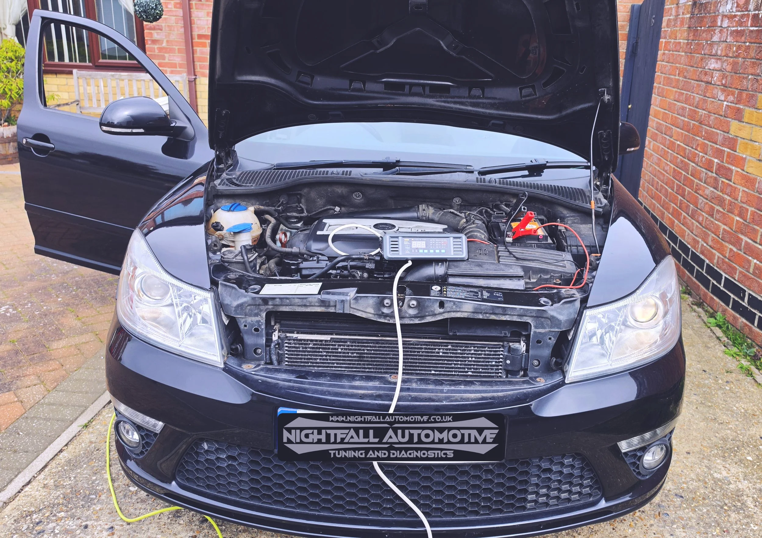 A black car with its hood open, showing the engine compartment and a diagnostic device connected to the engine. The car is parked outside next to a brick wall and a wooden fence.