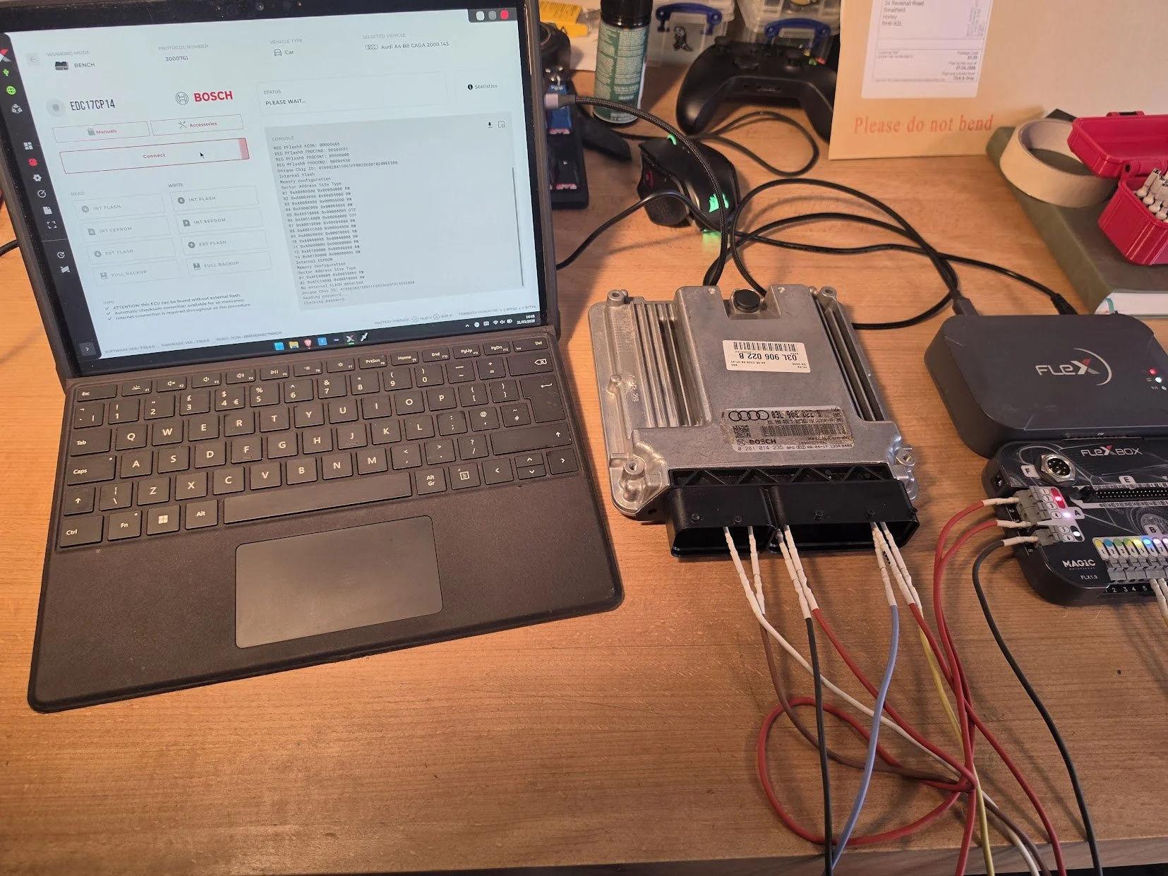 A workspace with a laptop displaying automotive diagnostic software, an automotive engine control unit (ECU) with connected wires, and a diagnostic interface device, all on a wooden table.