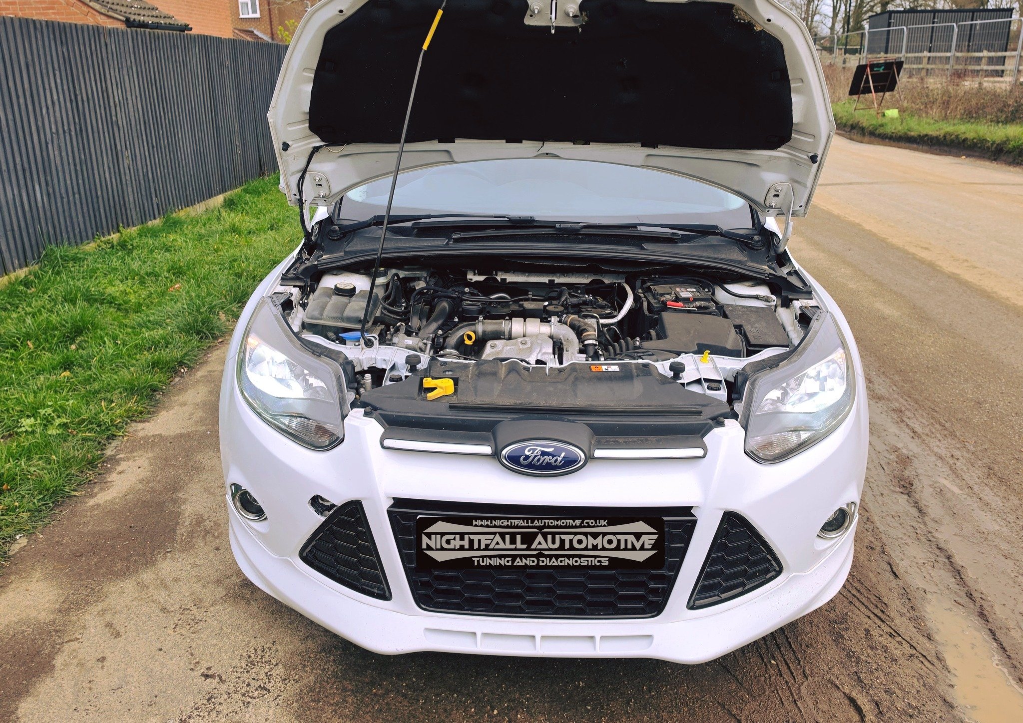 Front view of a white Ford car with open hood, showing the engine bay and various automotive components. The car has a black grille with a Ford logo and a license plate that reads 'Nightfall Automotive Tuning and Diagnostics.' The car is parked on a 