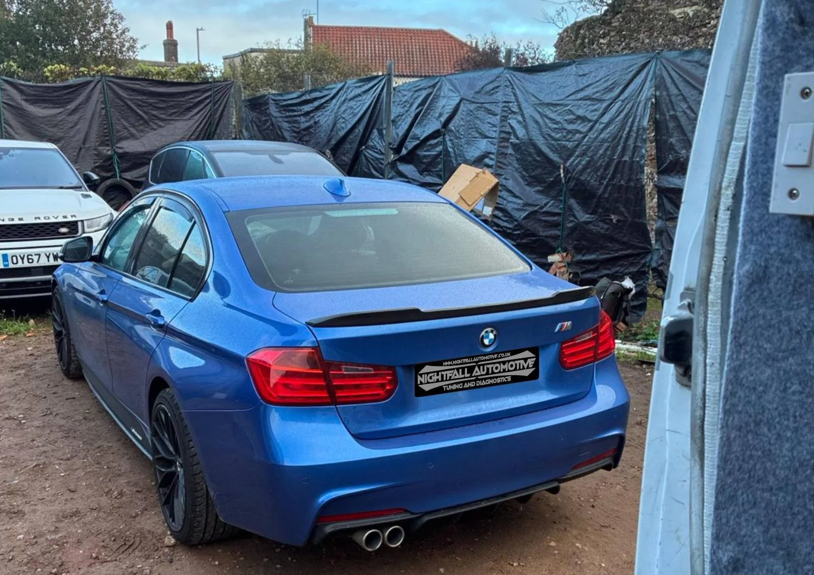 Blue BMW M3 parked on dirt ground at car repair shop, with other vehicles and black tarps in the background.