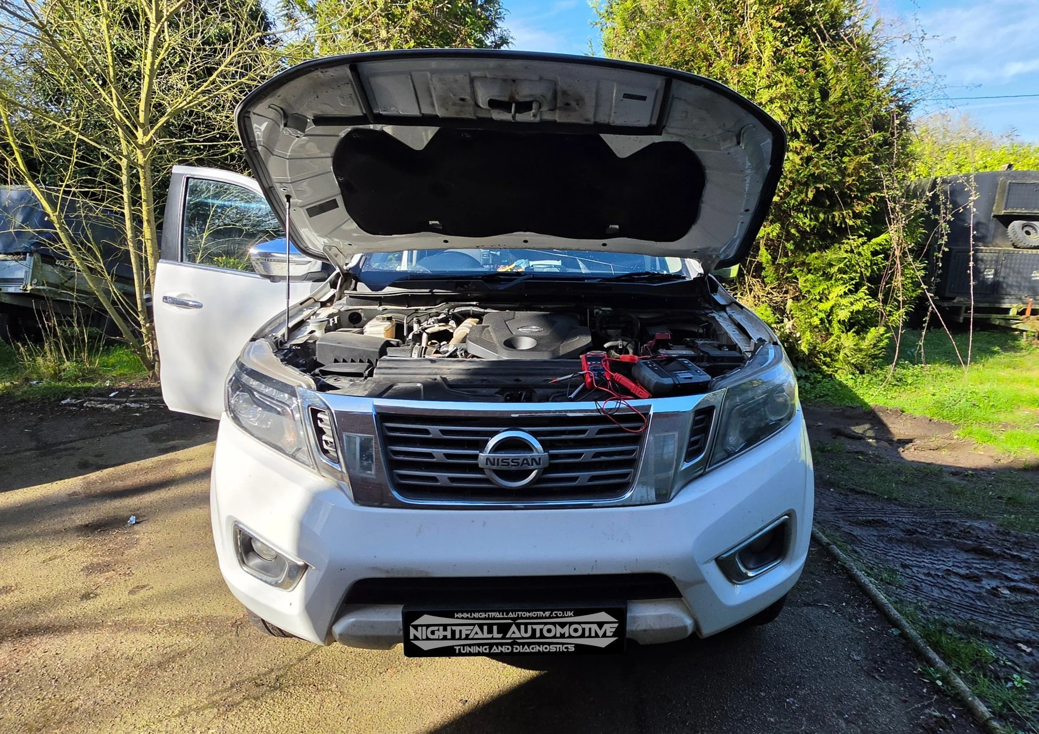 White Nissan SUV with open hood and red jumper cables attached to the engine, parked on a dirt surface in front of green shrubbery and trees.