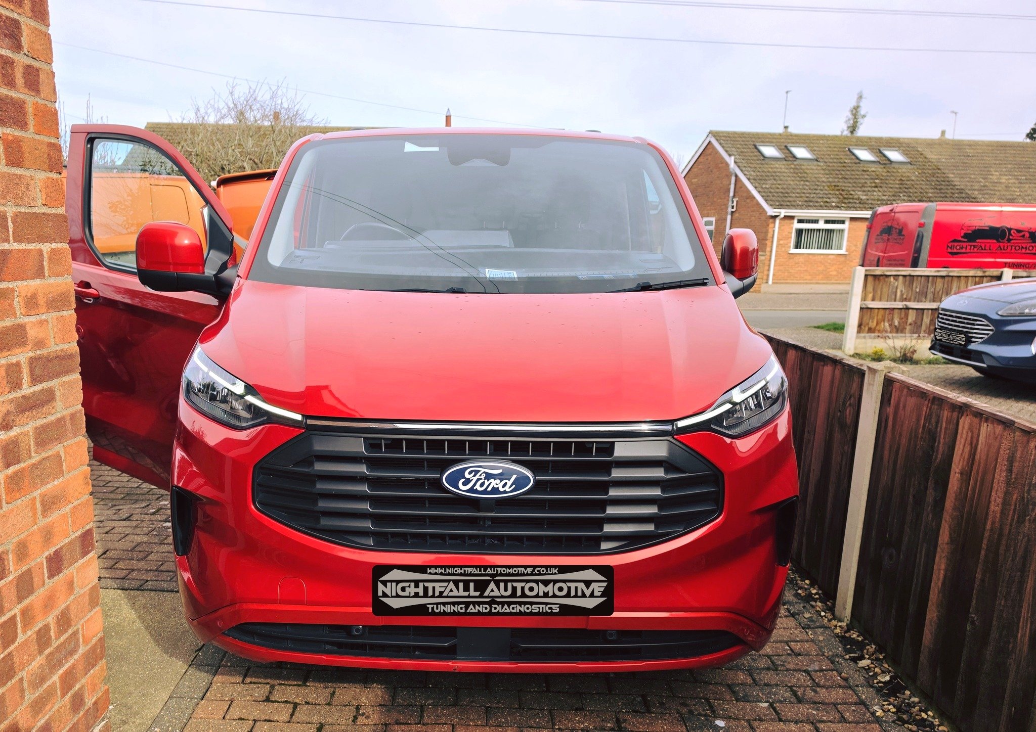 Red Ford vehicle parked next to a brick wall and wooden fence, with its driver's side door open, in a residential area.