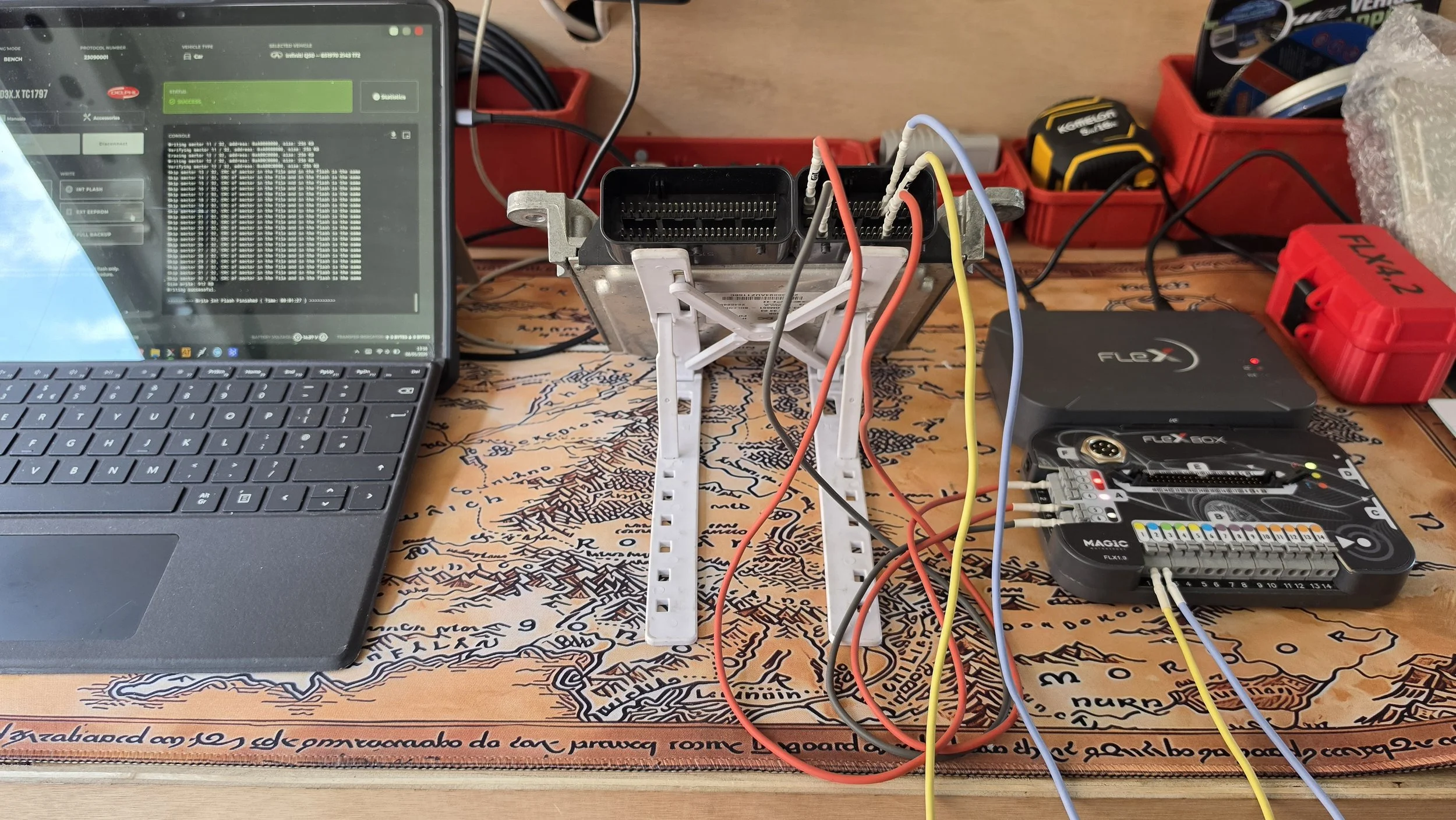 A computer setup on a table with electronic components, including a laptop, a circuit board, connected wires, and tools, indicating an electronic or hardware project in progress.