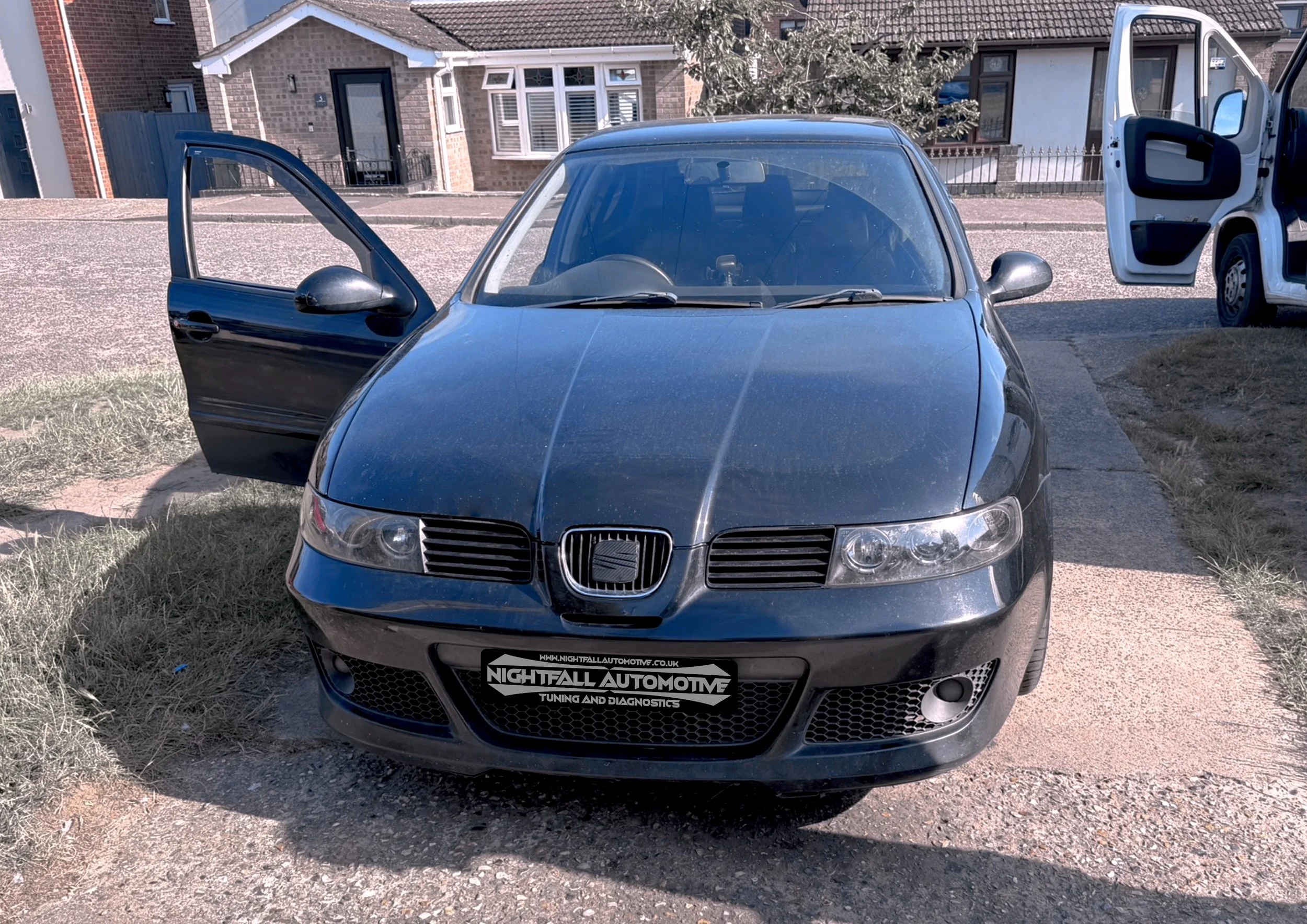 A gray car parked on a driveway with the front facing forward and the driver side door open, in front of residential houses.