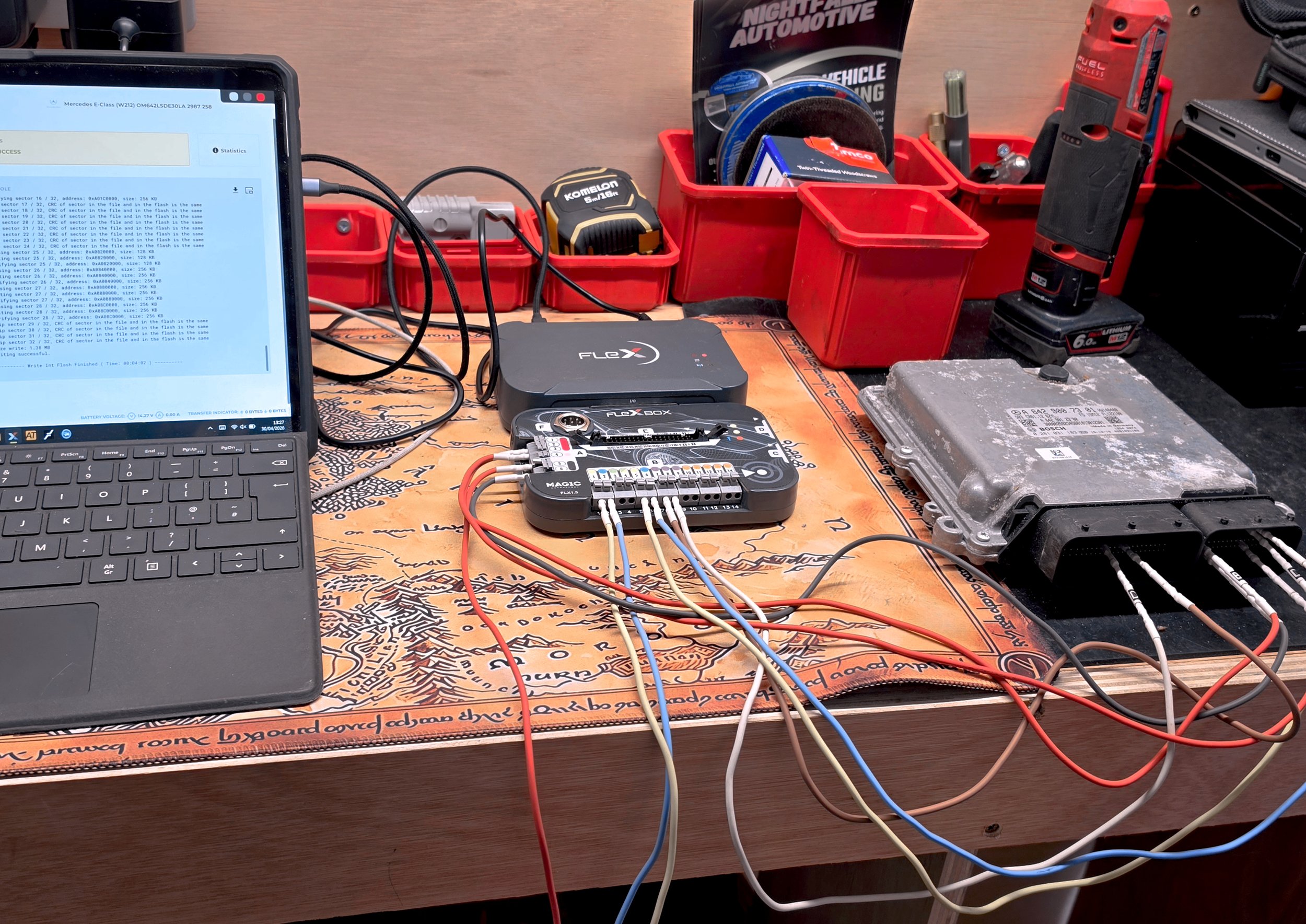 A workbench with automotive diagnostic tools, wires, a laptop, and a car engine control module.