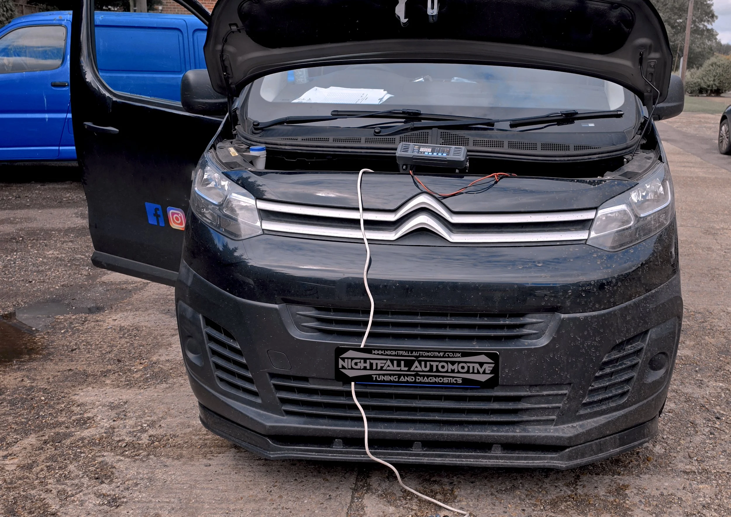 Front view of a black Citroën car with its hood open, revealing a diagnostic device placed on the engine. A white cable connects the device to the car. A blue vehicle is parked next to it, and a grassy area with trees and a clear sky are visible in t