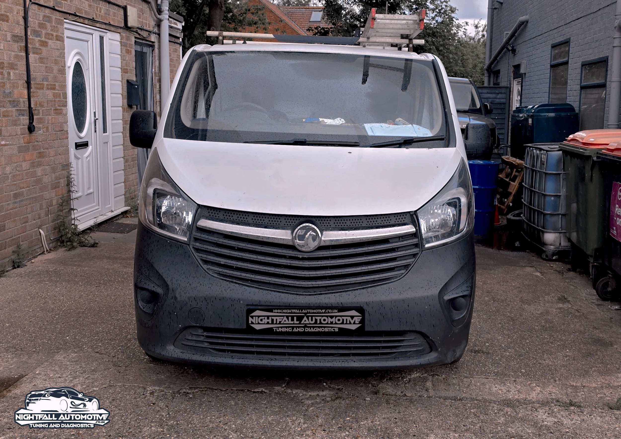 Front view of a white van parked in a residential driveway, with a ladder on the roof, rain droplets on the windshield, and various trash bins to the side. tuned by Nightfall Automotive