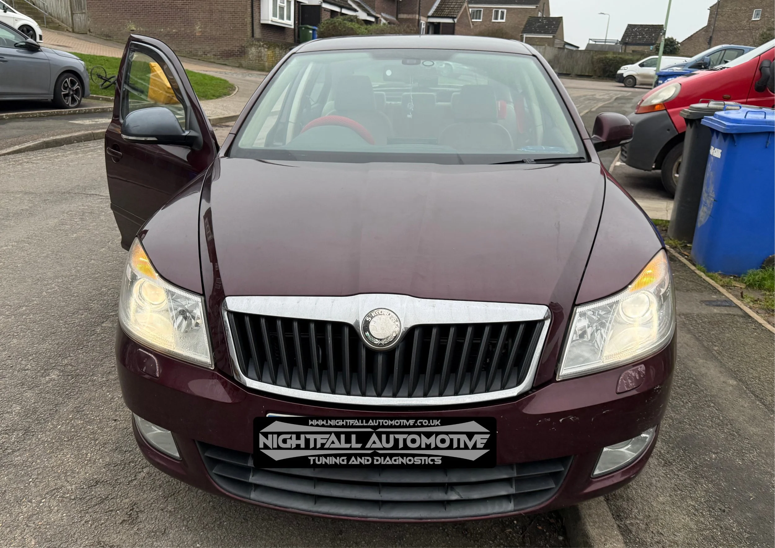 Front view of a maroon Skoda car with the driver’s side door open, parked in a residential area with other cars and houses visible in the background.