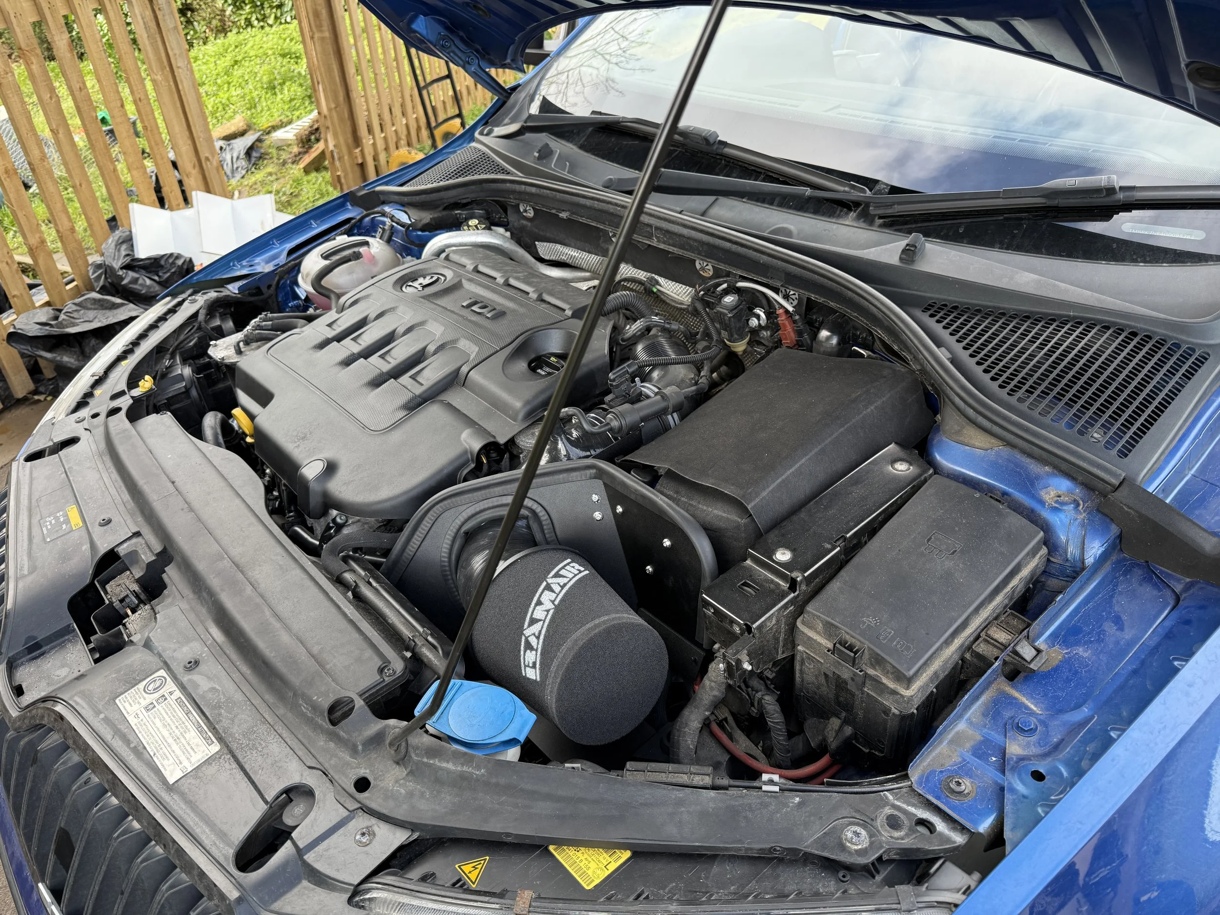 Under the hood of a blue car showing the engine and various components, including a RAM air filter, battery, and fluid reservoirs.