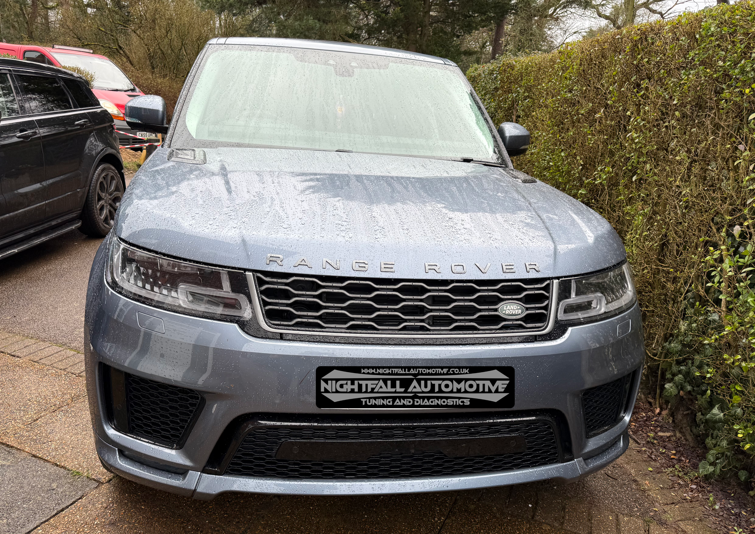 A gray Range Rover SUV parked outdoors on a wet pavement with raindrops on its surface, next to a hedge and other parked cars.