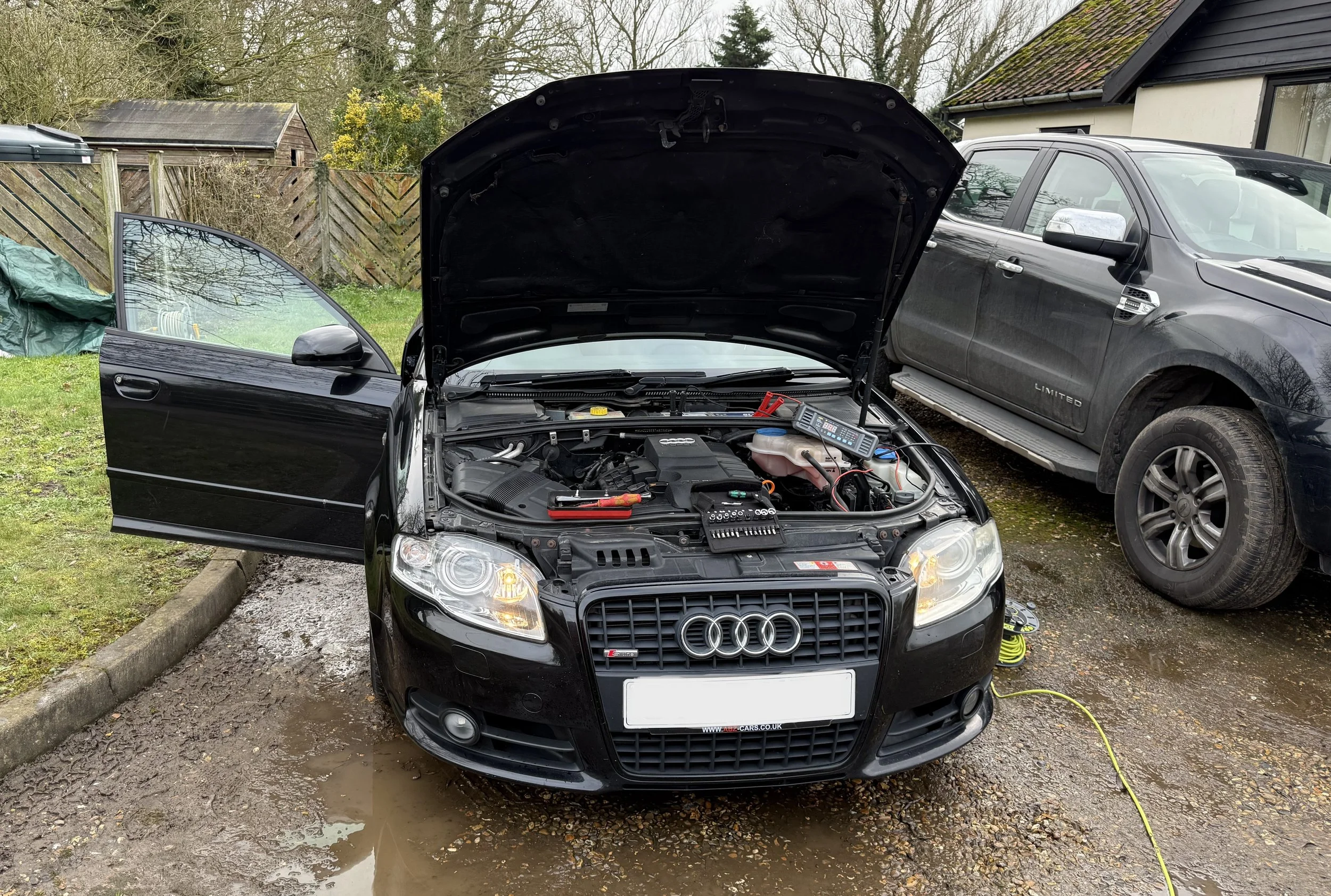 A black Audi sedan with its hood open, showing the engine, parked next to a black pickup truck on a muddy driveway. The driver's side door of the Audi is open, and a diagnostic scanner and jumper cables are on the engine.