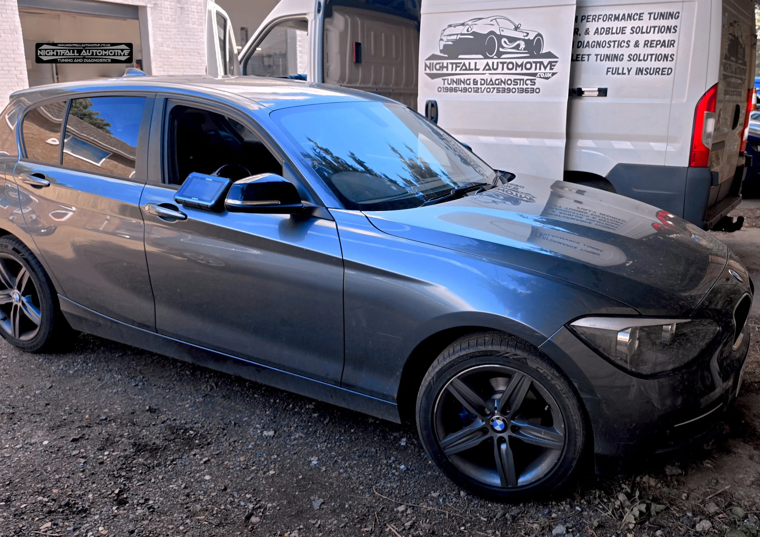 A shiny gray BMW parked in front of a white vehicle with advertising for Nightfall Automotive, an automotive tuning and diagnostics company, on the side. tuned by Nightfall Automotive