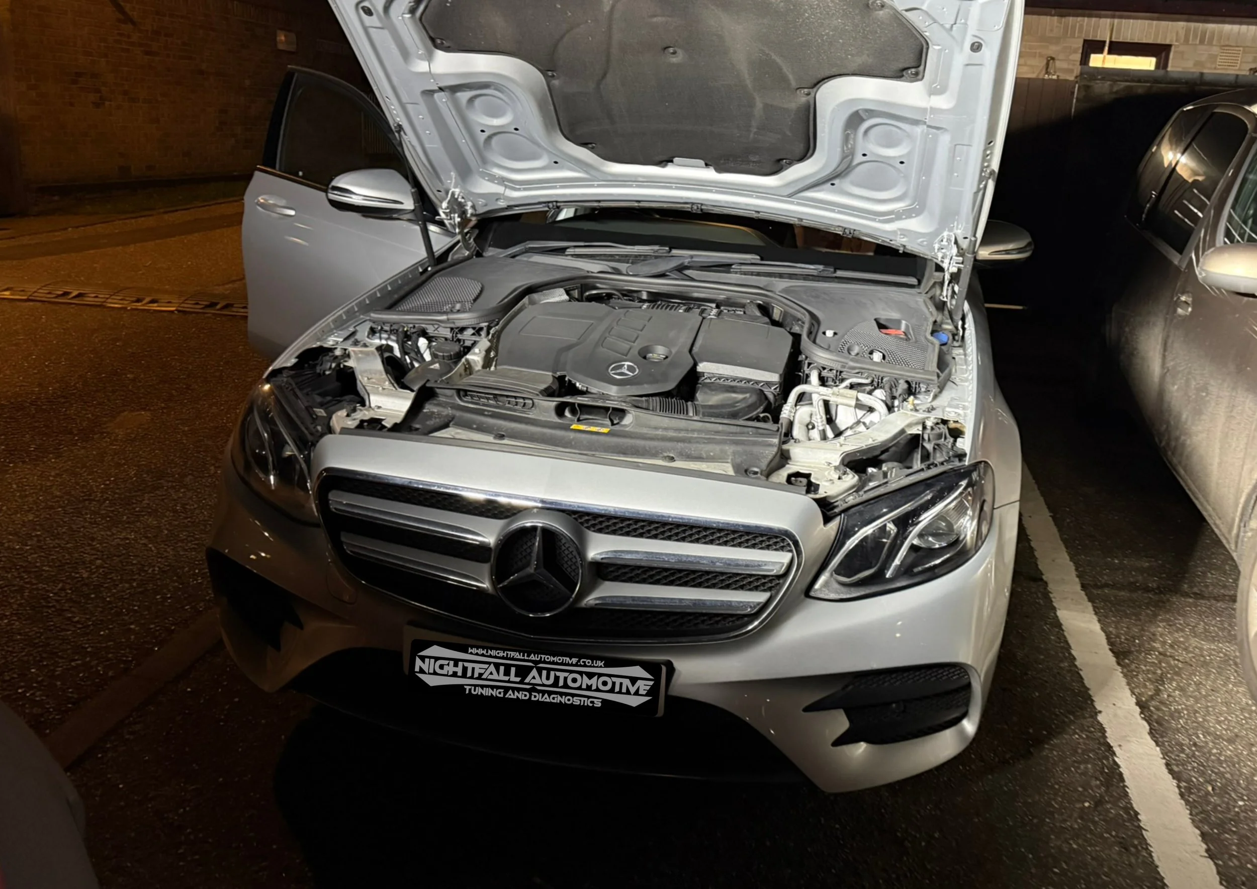A silver Mercedes-Benz car with its hood open, showing the engine. The car is parked in a parking lot at night, next to a gray vehicle. The front license plate reads 'Nightfall Automotive Tuning and Diagnostics'.