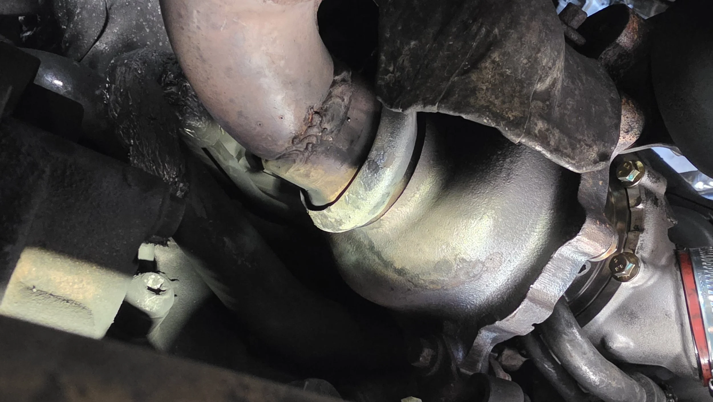 Close-up of engine parts, showing a rusty exhaust manifold, metal pipes, and bolts. Tuned by nightfallautomotive