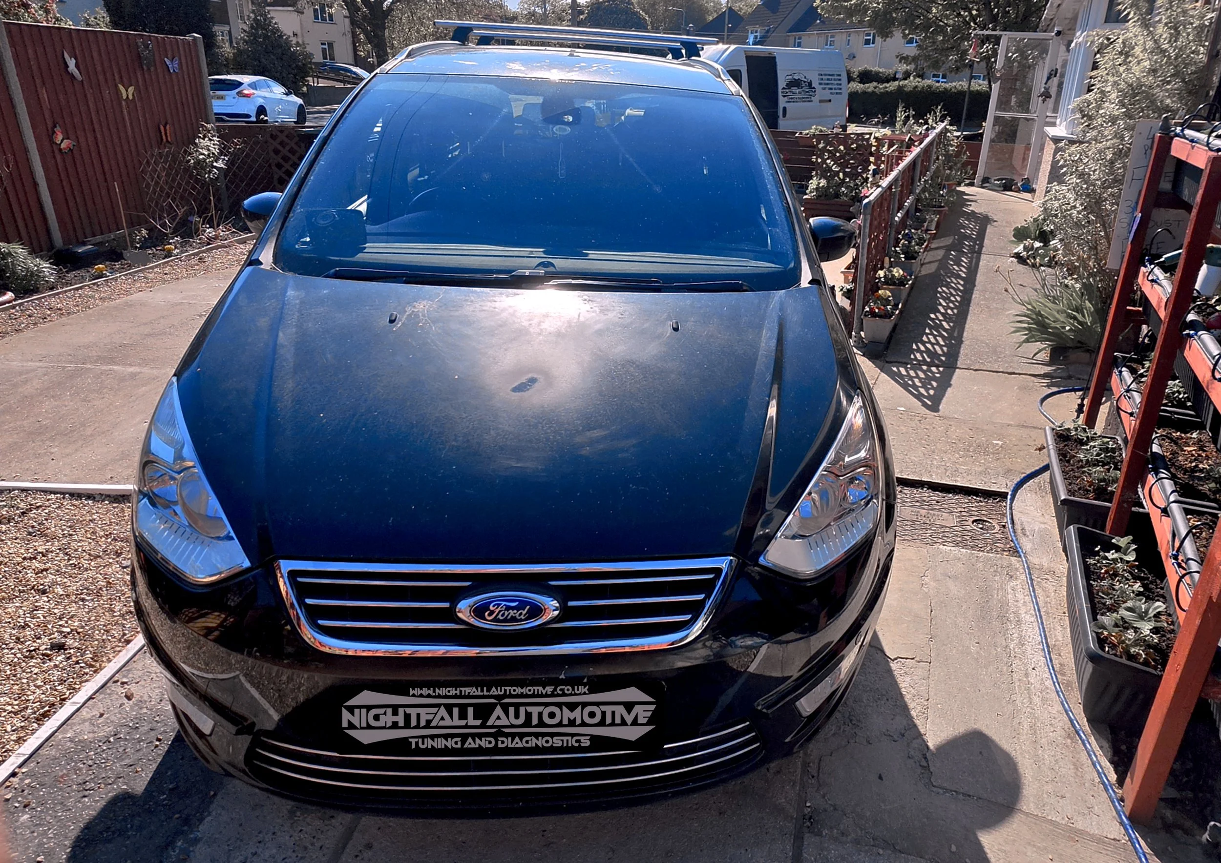 A black Ford car parked in a driveway with a blue roof rack on top, surrounded by a garden and residential buildings in the background.