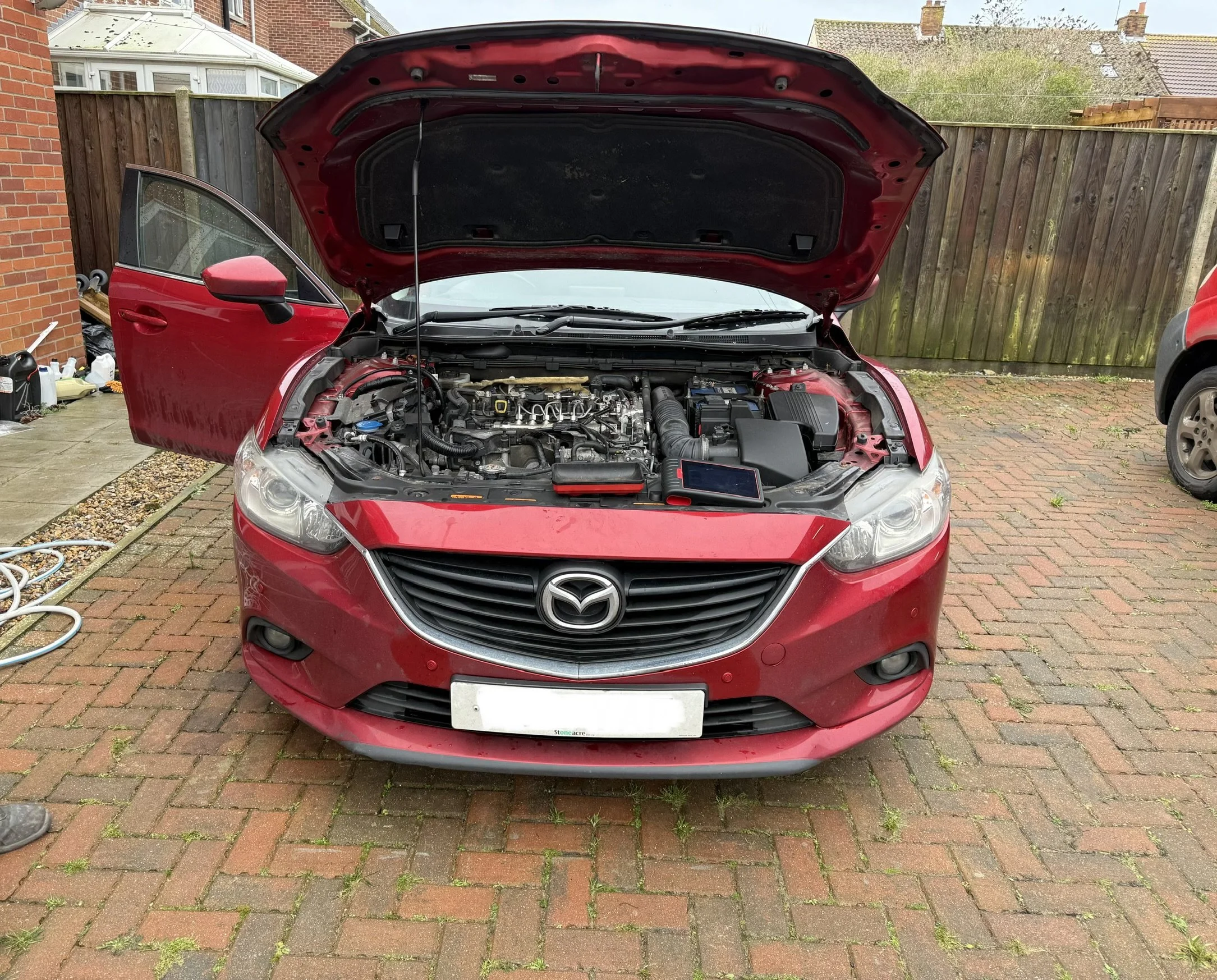 Red Mazda car with open hood showing engine, parked on brick driveway with open driver side door, in a residential area. Stage 1 sorted By Nightfall Automotive